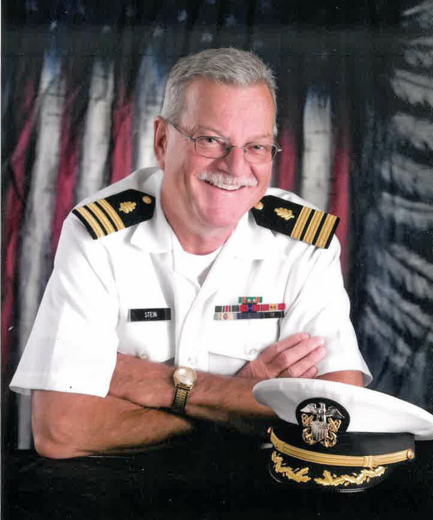 Smiling gray-haired naval officer in dress whites with ribbons, sitting at a desk, glasses on, a decorated cap resting nearby on the table.
