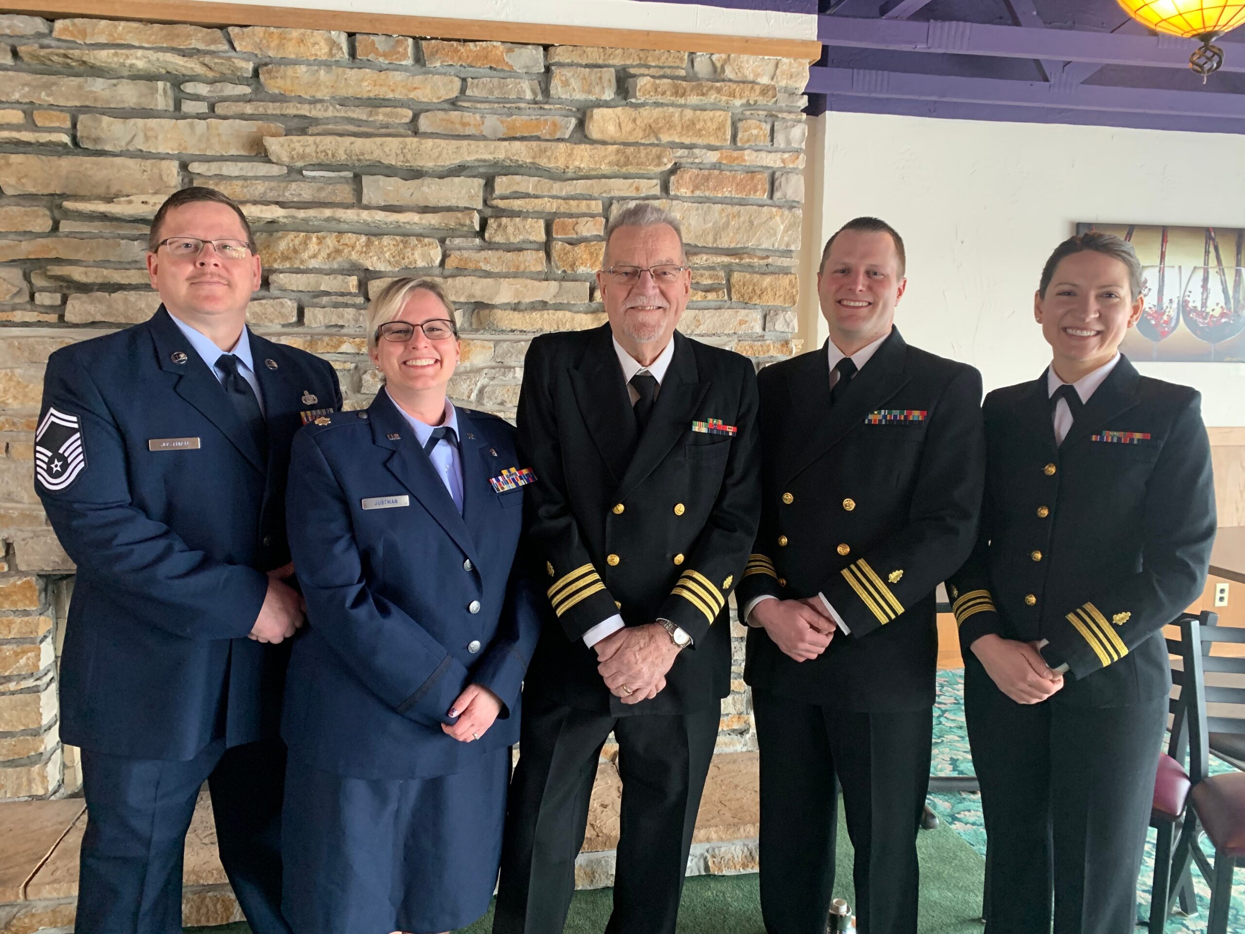 Five people in dress uniform posing indoors in front of a stone wall, smiling at the camera.