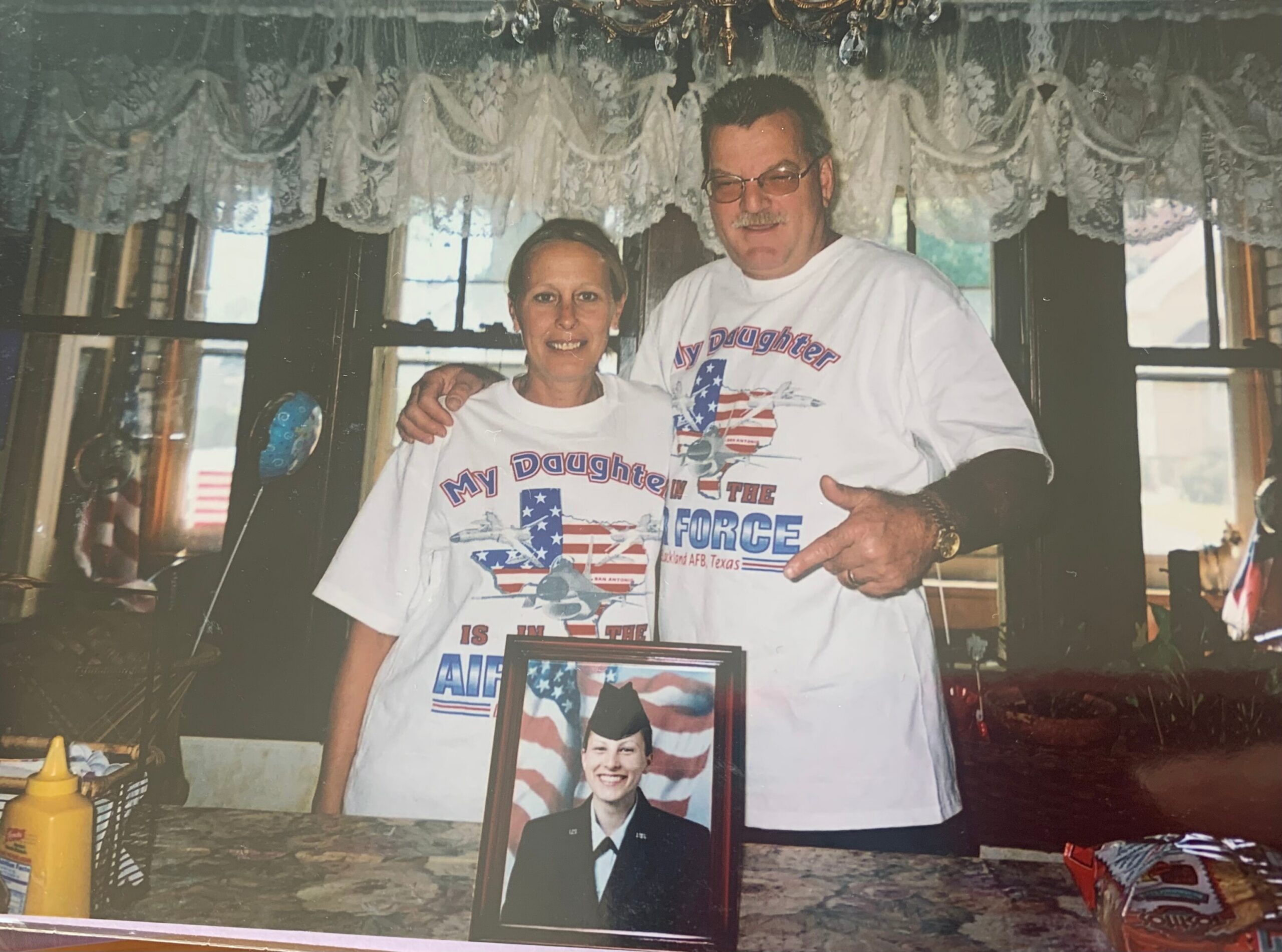 A couple wearing matching 'My Daughter is in the Air Force' shirts pose indoors, with a framed portrait on a table in front of them.