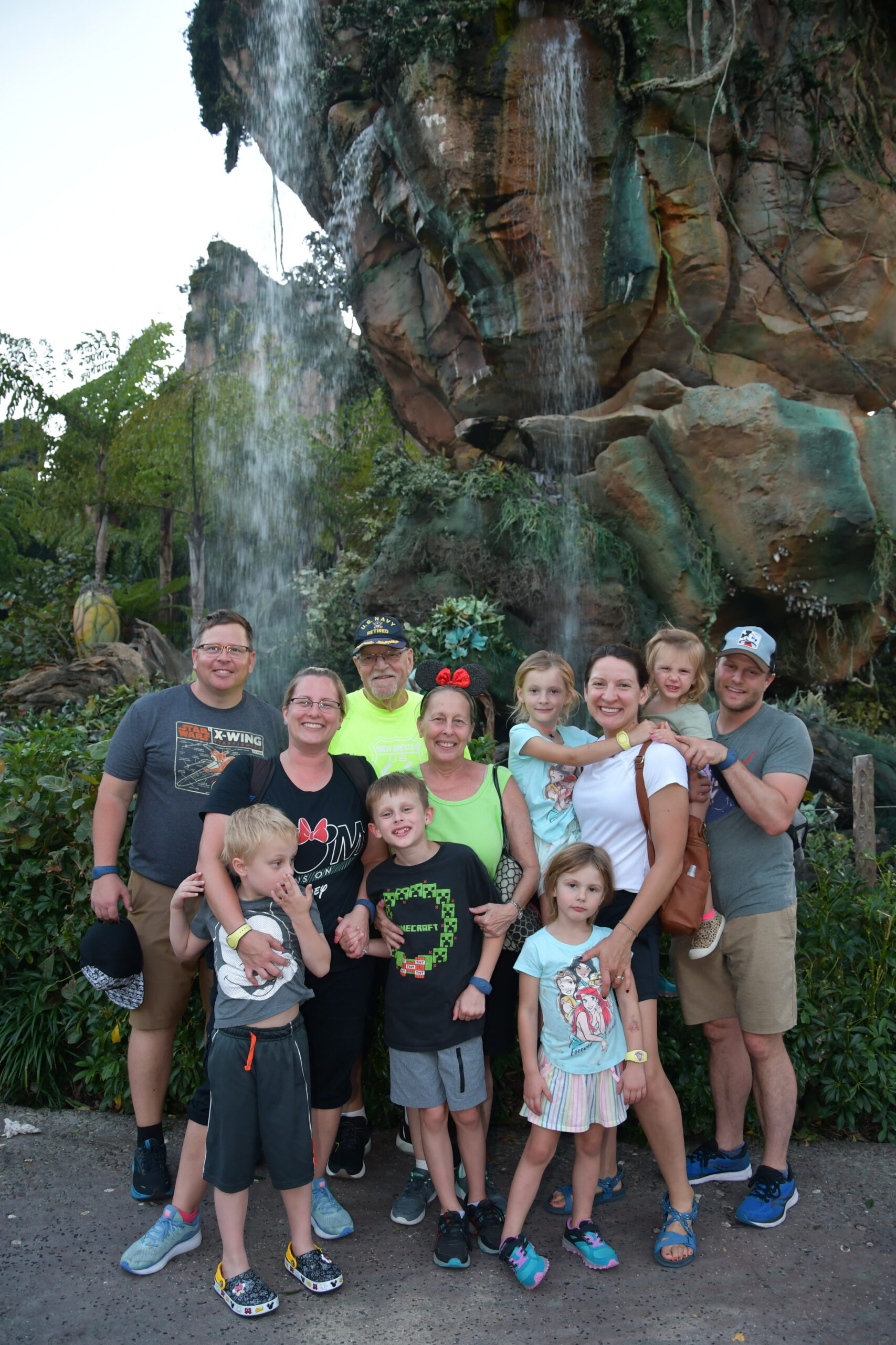 A large group of people posing together outdoors in front of a rocky waterfall and greenery, including adults and children smiling at the camera.