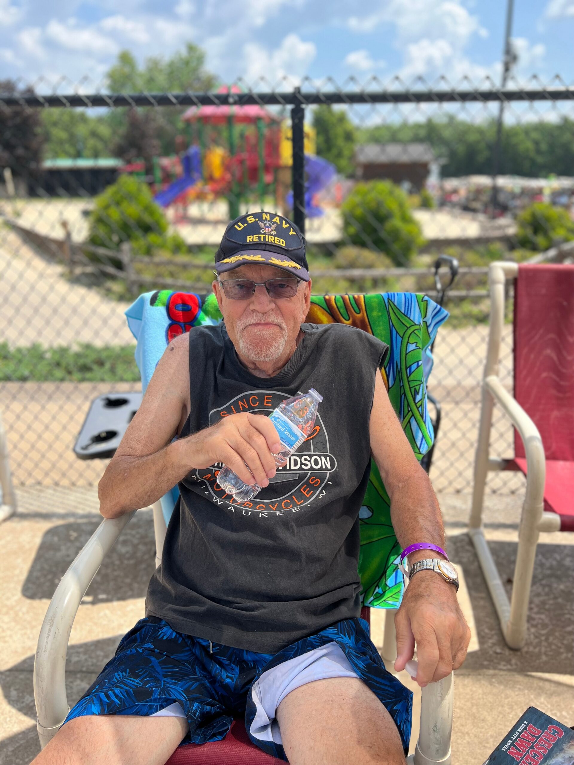 Older man wearing a U.S. Navy Retired cap sits in a folding chair outdoors, holding a water bottle.