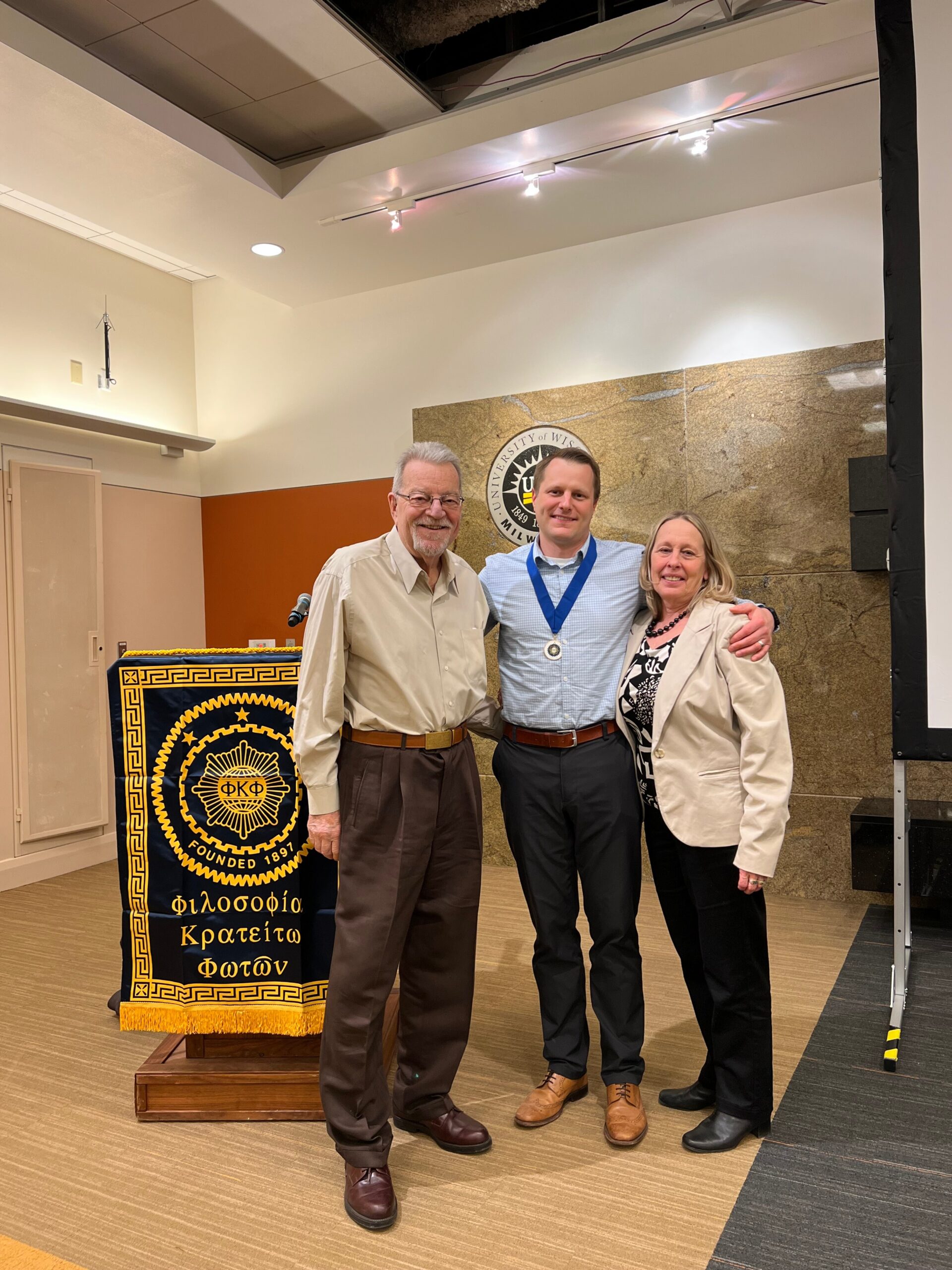 Three adults pose together at an award ceremony, the man in the center wears a medal on a blue ribbon and smiles beside two colleagues. A Greek-language banner and emblem are visible in the background.