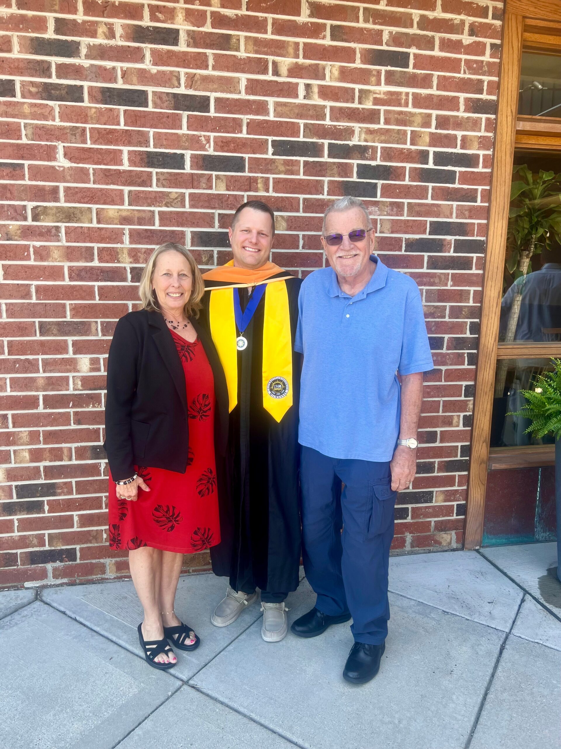 Graduate in black gown with yellow stole and medallion, flanked by a woman in red dress and a man in blue polo, posing outside a brick wall.
