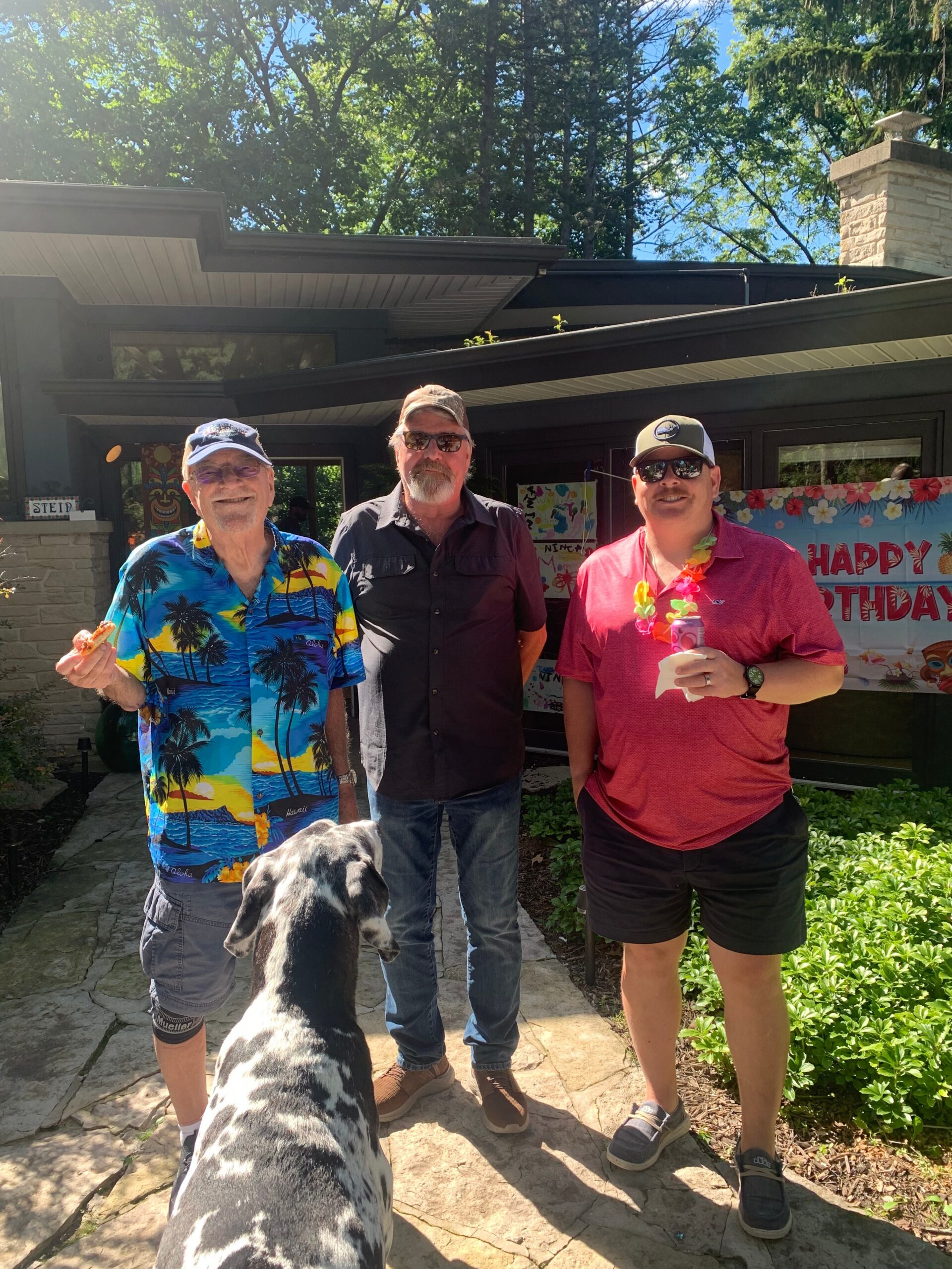 Three men pose outside a house for a birthday celebration, with a large spotted dog in the foreground.