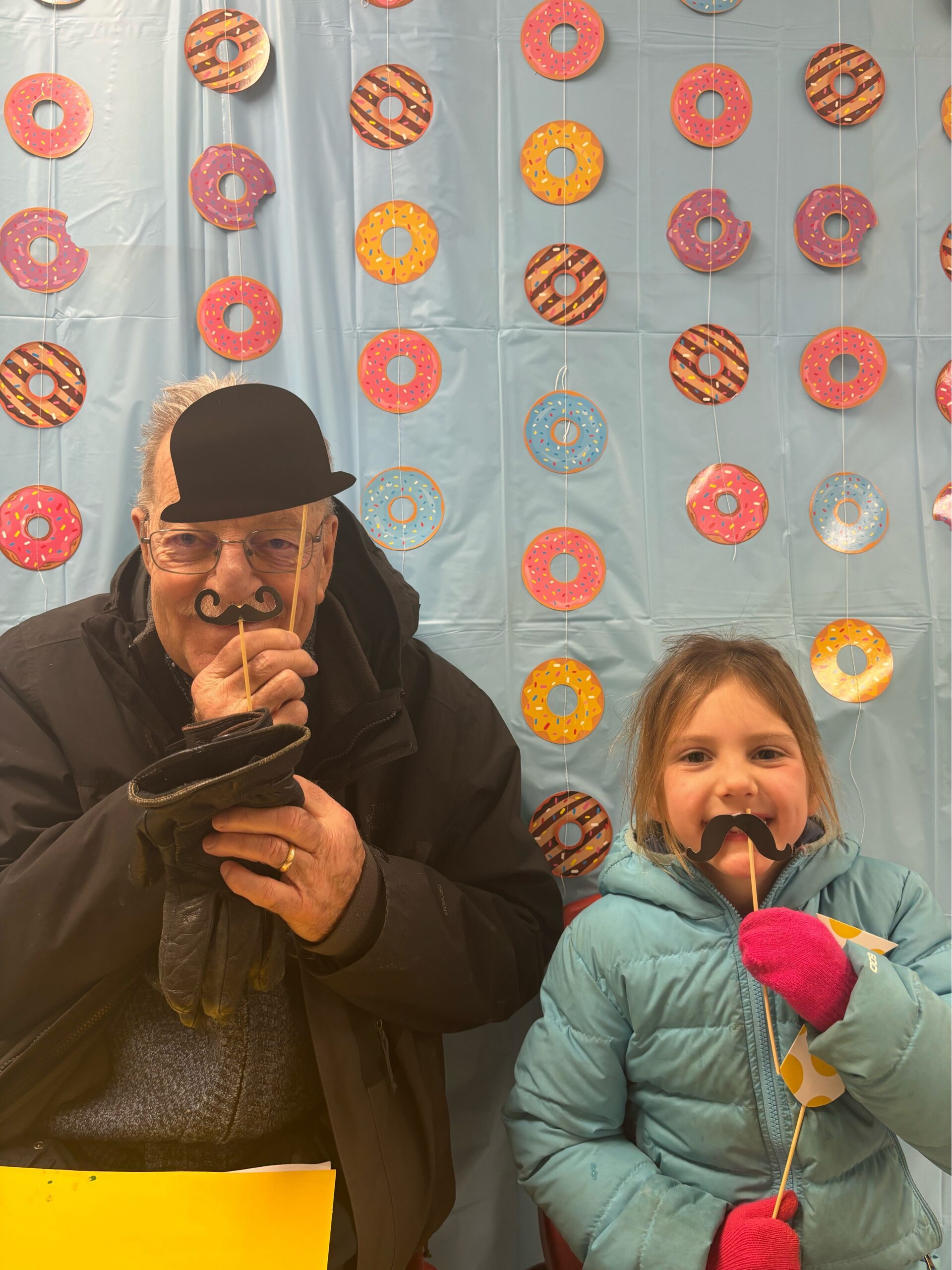 Elderly man and young girl posing with props in front of a donut-themed backdrop, holding stick-on mustache and hat props.