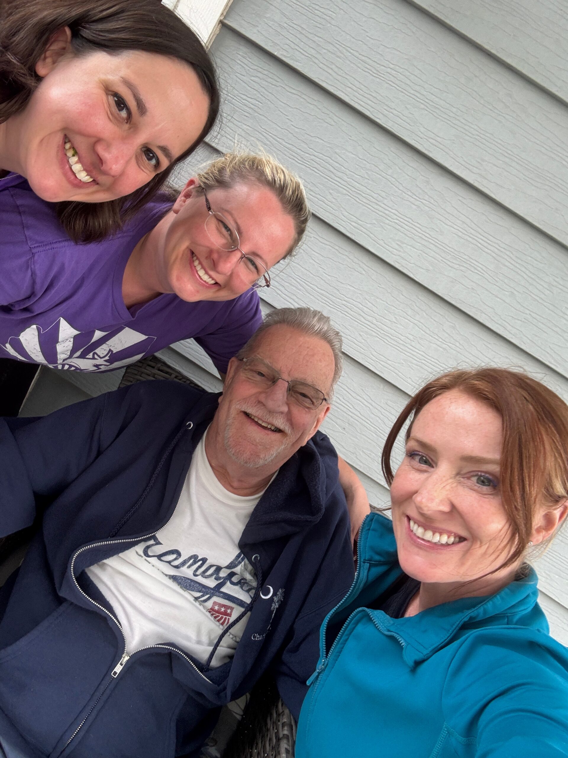 Four friends posing for a close-up outdoor selfie, smiles all around, against a gray wooden wall.