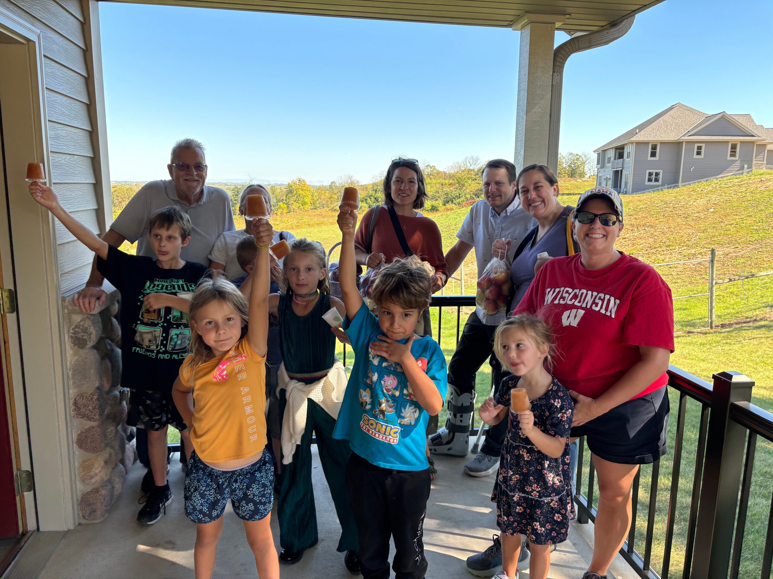 Smiling group of children and adults on a balcony, holding orange popsicles on a sunny day with a suburban landscape behind them.