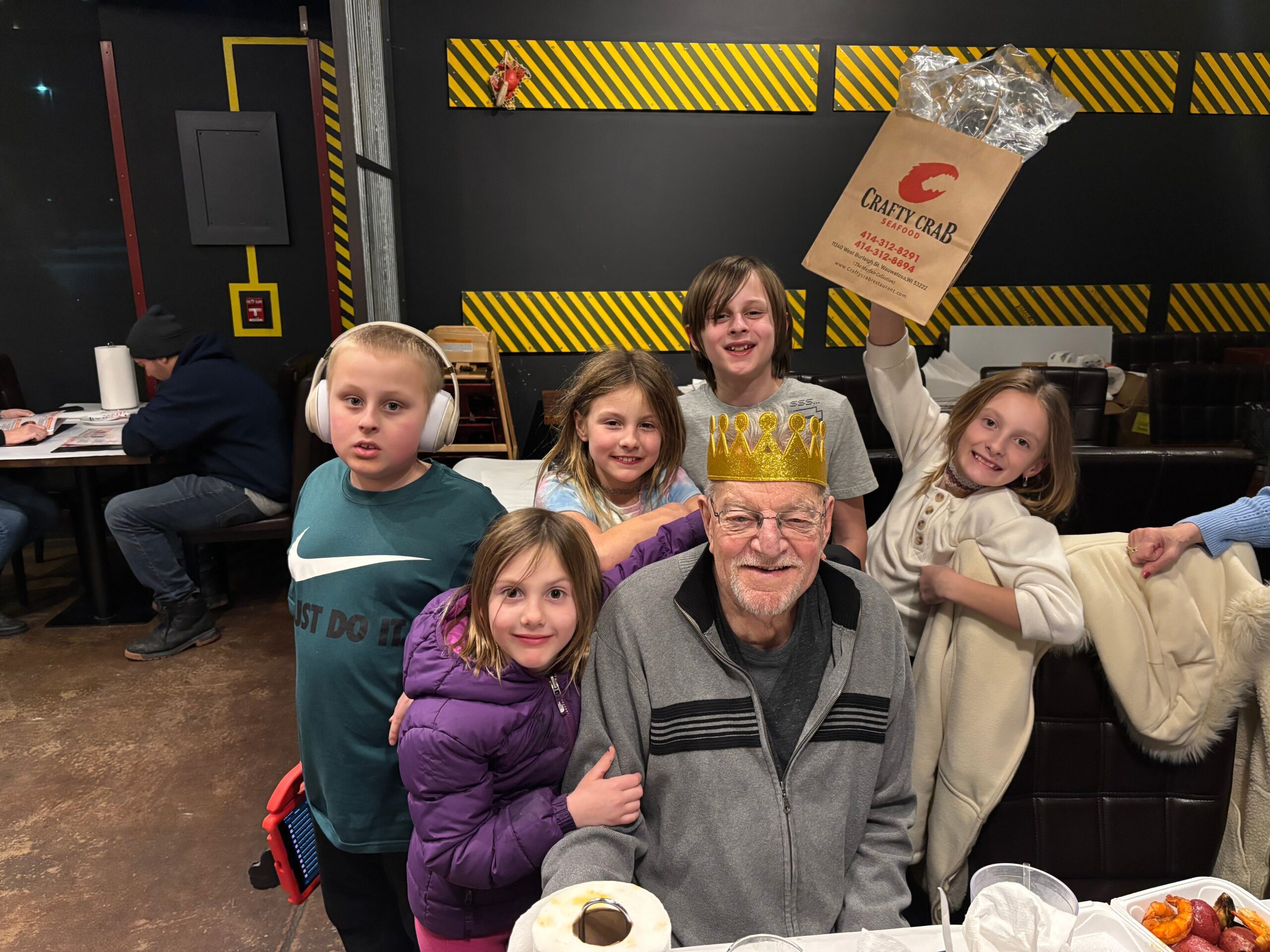 Group of smiling kids gathered around a table with snacks, an older man wearing a gold crown sits at the front in a party room with black walls and yellow striped safety tape in the background.
