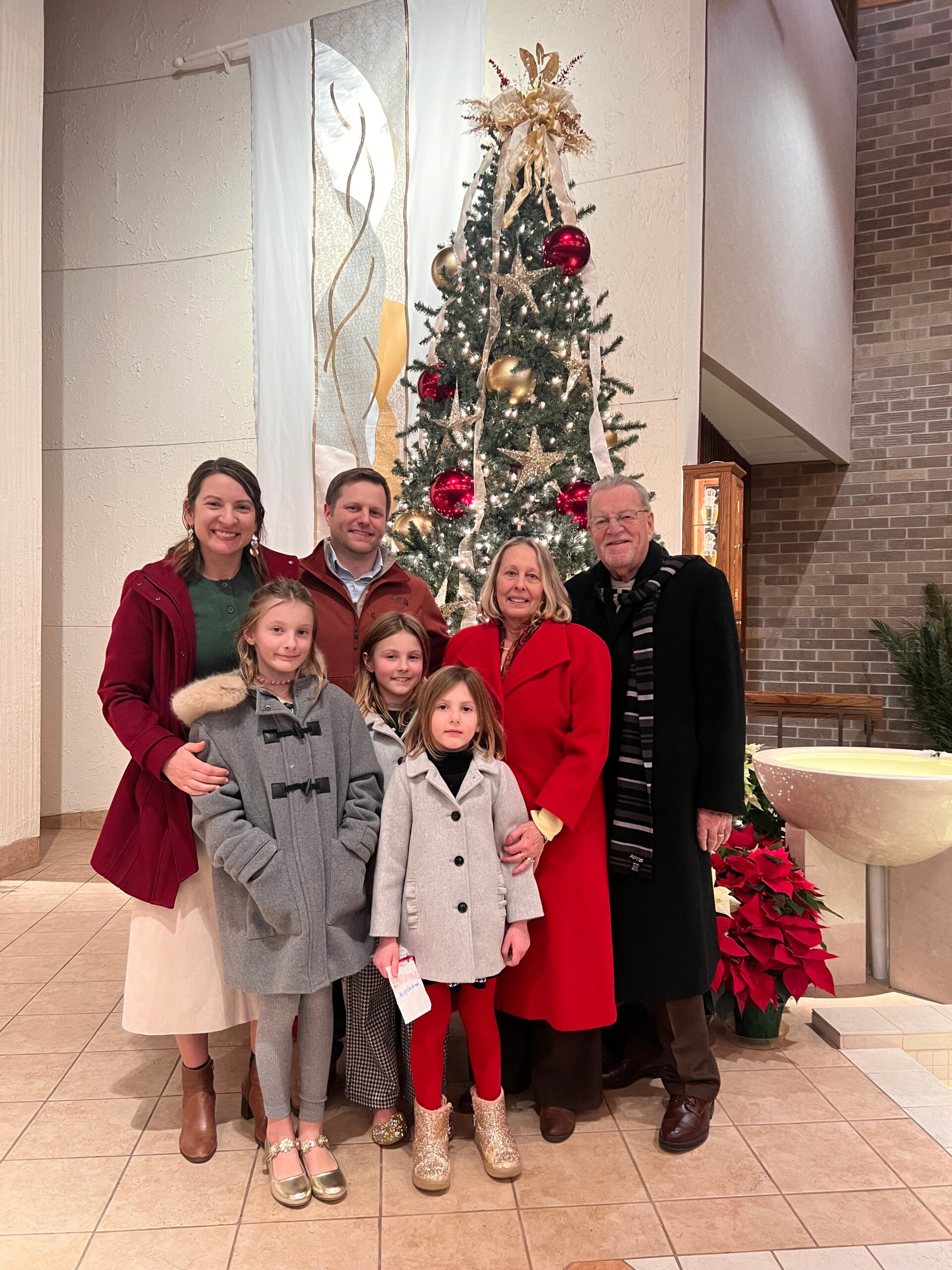 Family group posing in front of a decorated Christmas tree inside a building lobby, smiling at the camera.