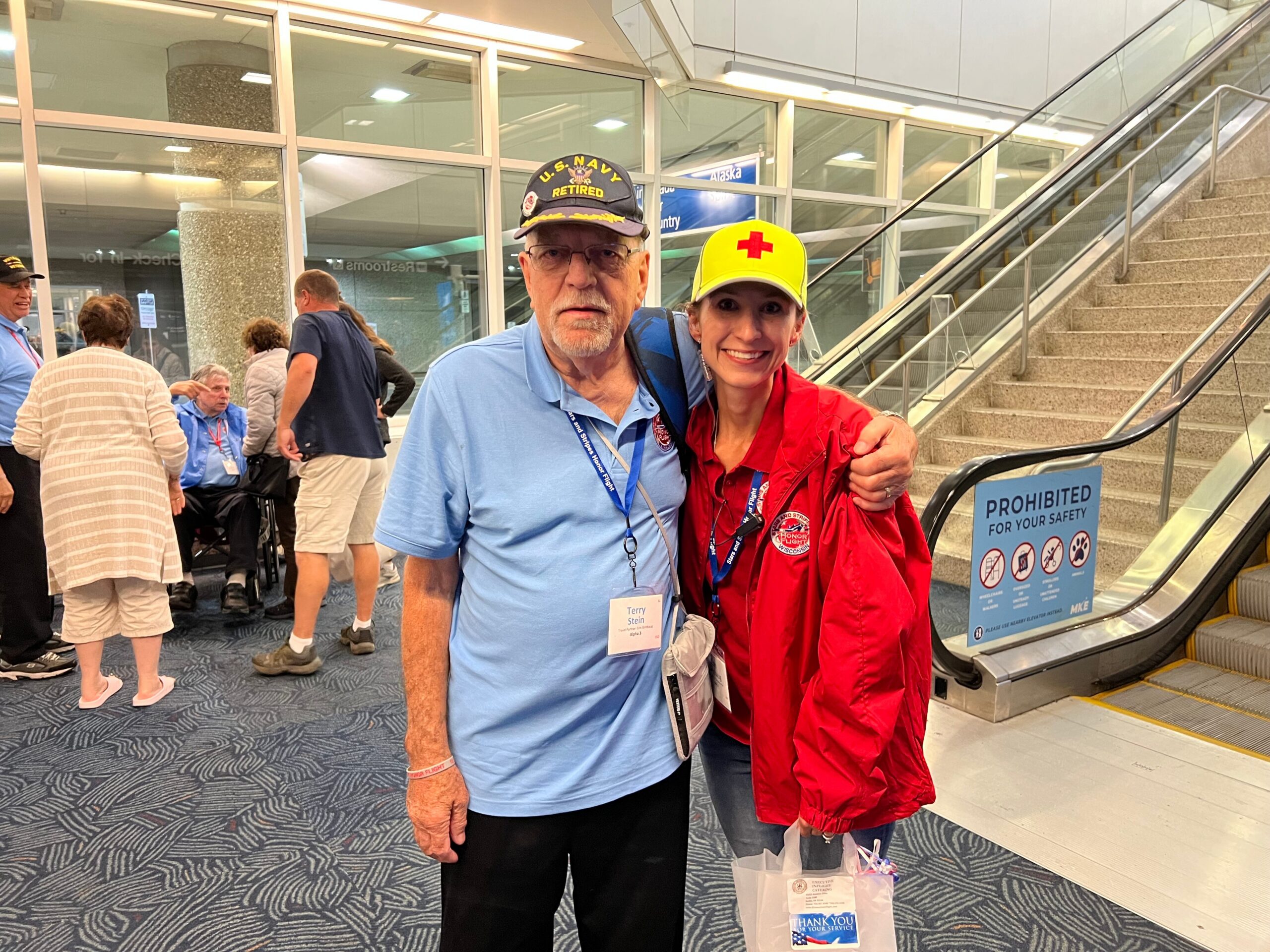 Smiling man in a light blue polo with a U.S. Navy Retired cap stands beside a woman in a red jacket and a yellow cap with a red cross in an airport terminal.