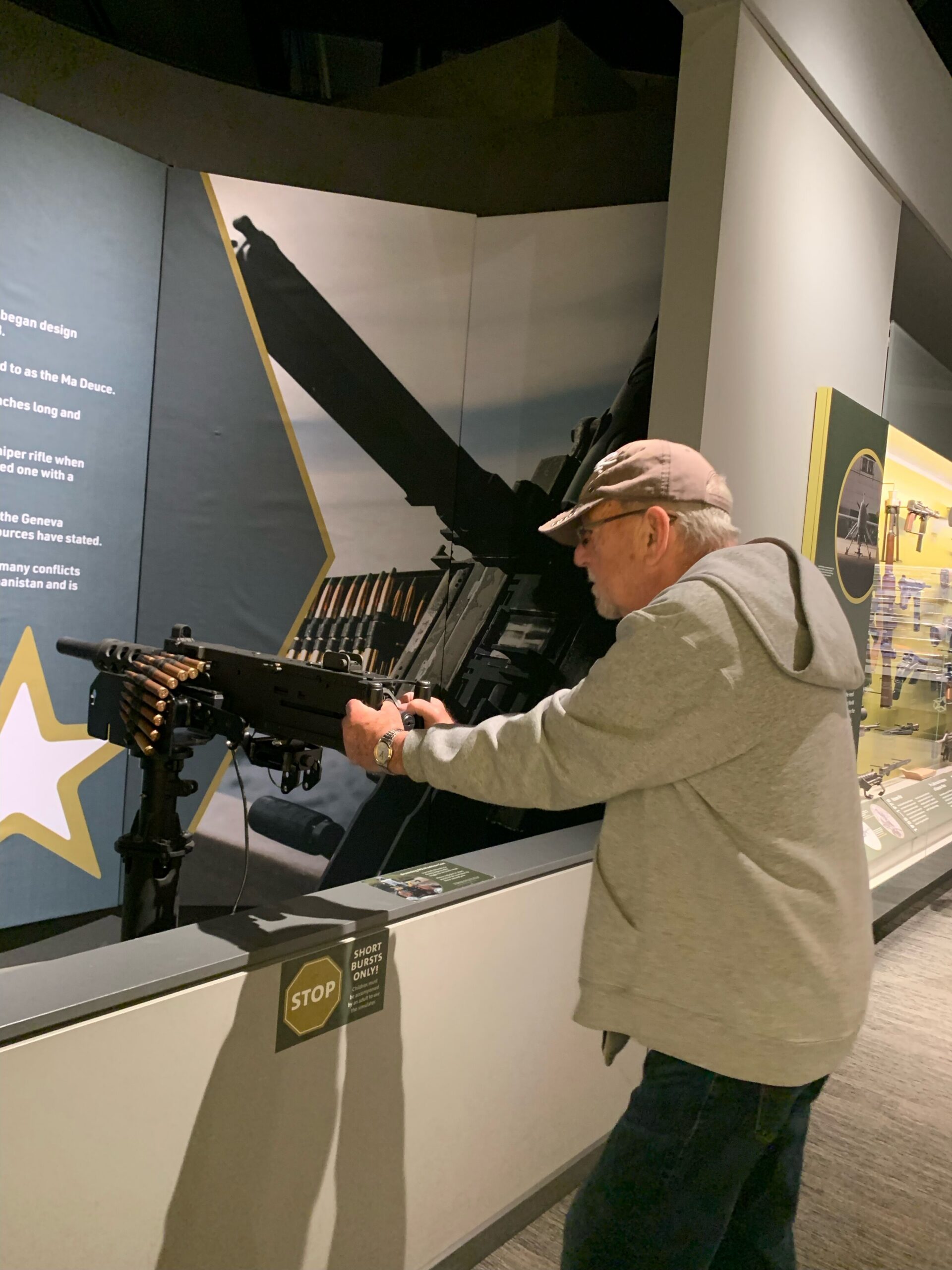Man in a gray hoodie and cap handling a large mounted rifle in a museum exhibit.