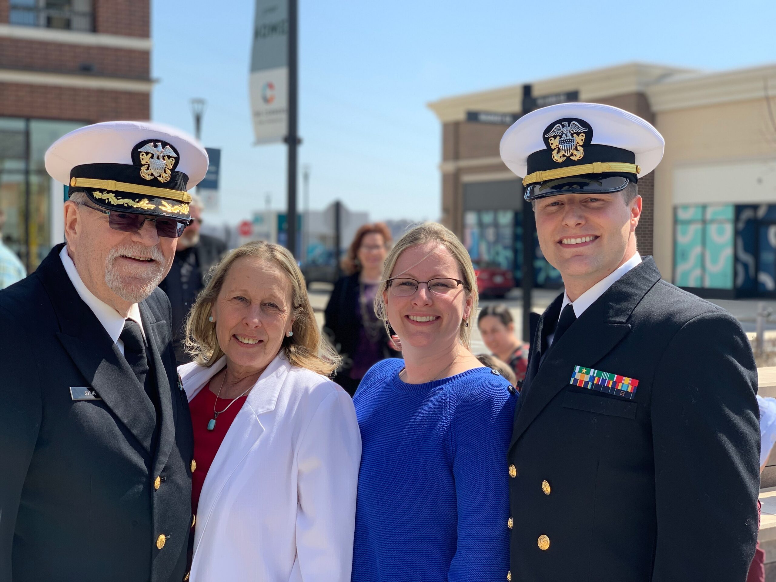 Group of smiling people taking a sunny outdoor selfie; two men in formal military dress uniforms with hats and medals, plus a woman in a blue sweater and others partially visible