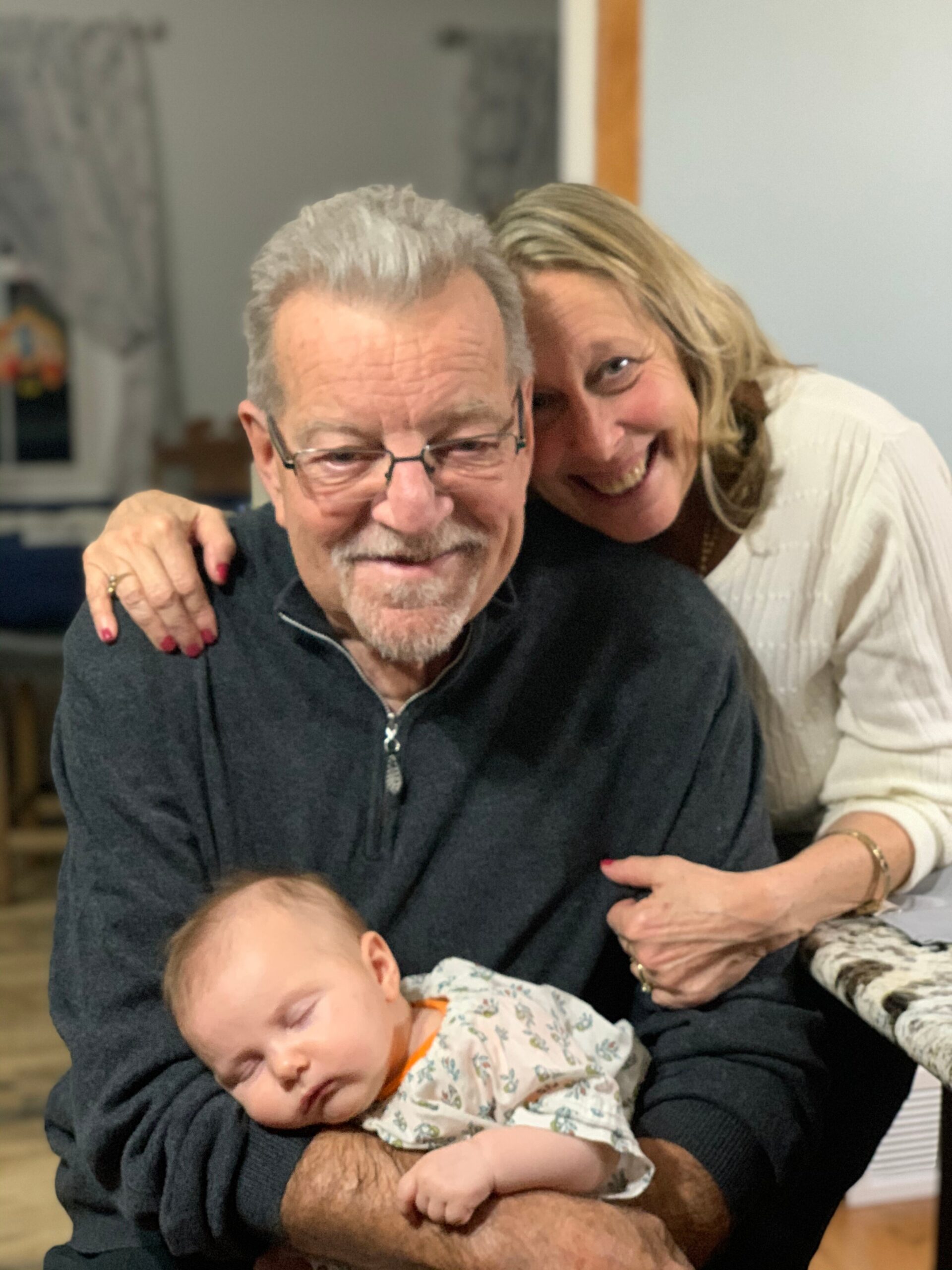 Older man with glasses holding a sleeping baby, with a smiling woman leaning in from behind.