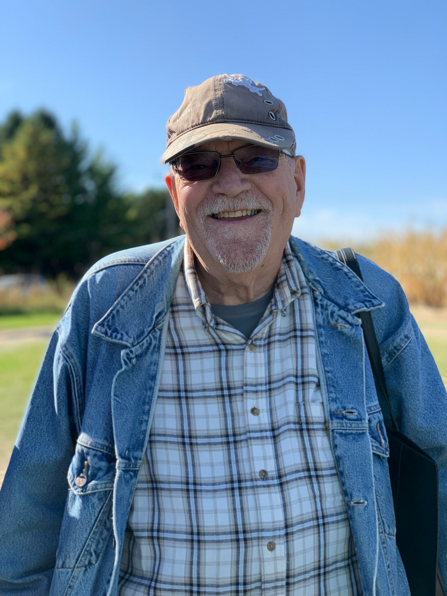 Smiling older man in a denim jacket and plaid shirt outdoors, wearing a gray cap and sunglasses against a clear blue sky.