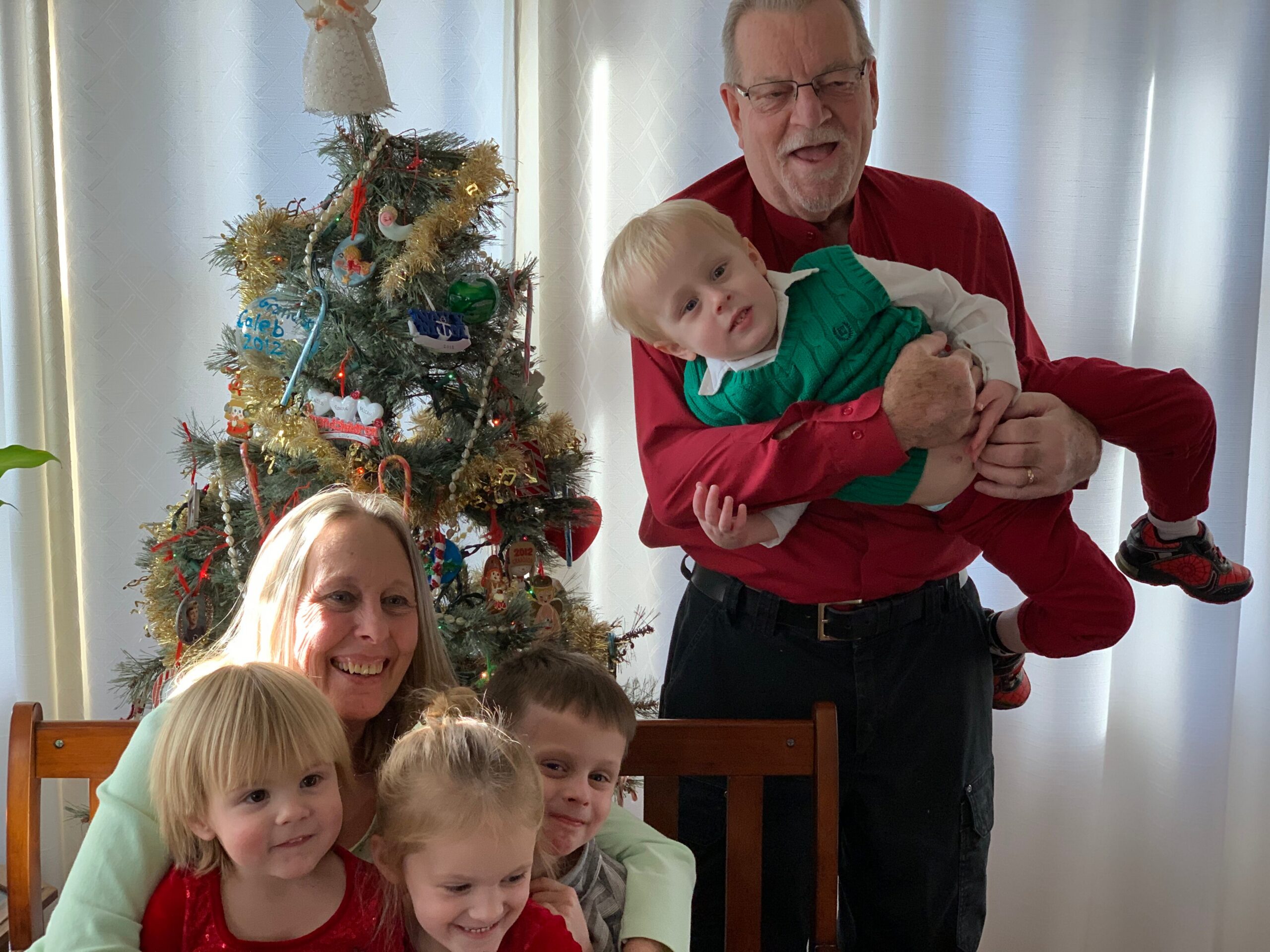 Family posing by a decorated Christmas tree, smiling children in foreground and an adult man in red holding a young boy.