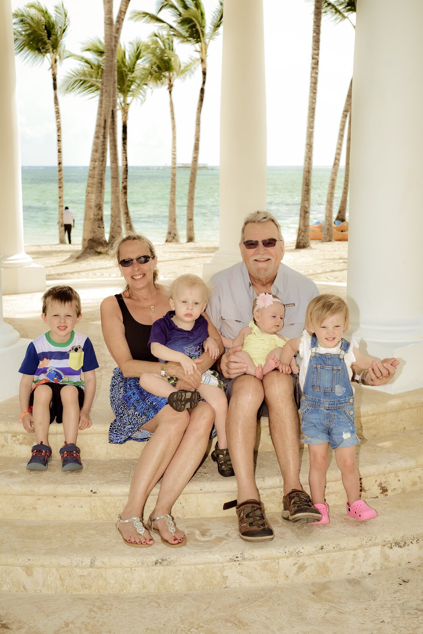Multigenerational family posing on stone steps at a sunny beach with palm trees and the ocean in the background.