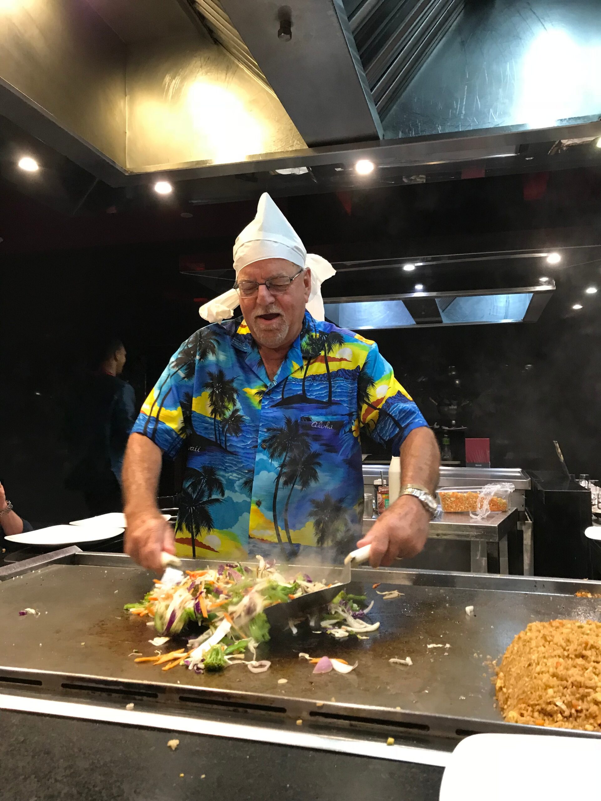 Chef wearing a white hat and colorful tropical shirt cooks at a flat-top grill, tossing vegetables on a hibachi-style station in a restaurant kitchen.