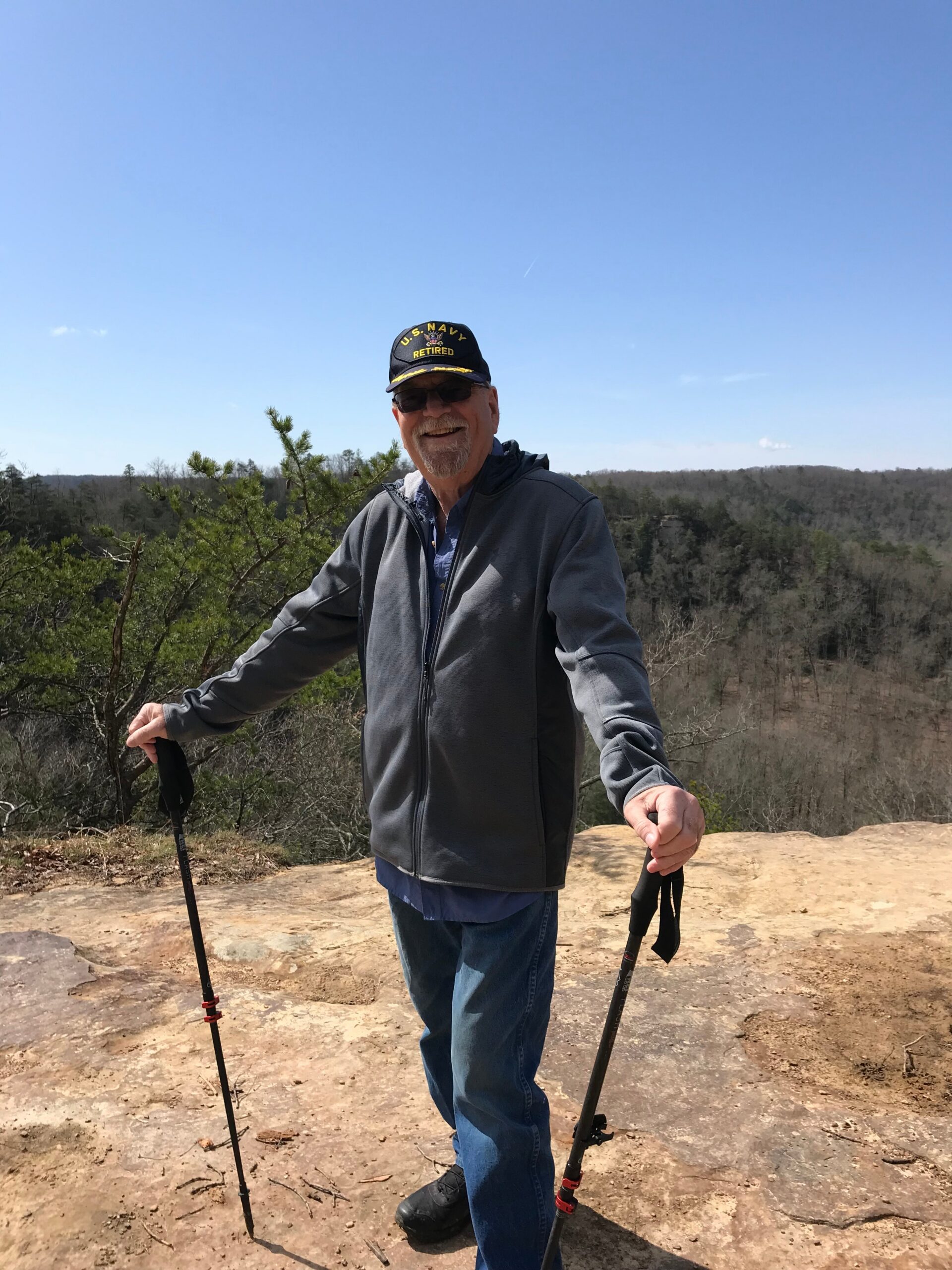 Smiling older man in a gray jacket and U.S. Navy Retired cap stands on a rocky overlook, holding trekking poles against a forested valley and blue sky.