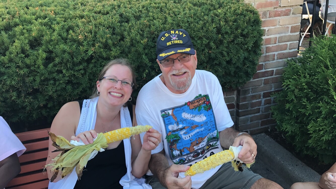 Smiling woman and man on a bench outdoors, each holding corn on the cob with husks in view, green hedges behind them.