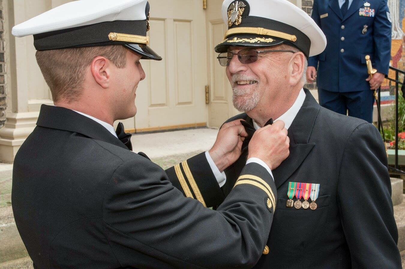 Two military officers in dress uniforms; a young man adjusts an older man's bow tie, medals visible on his chest outdoors.