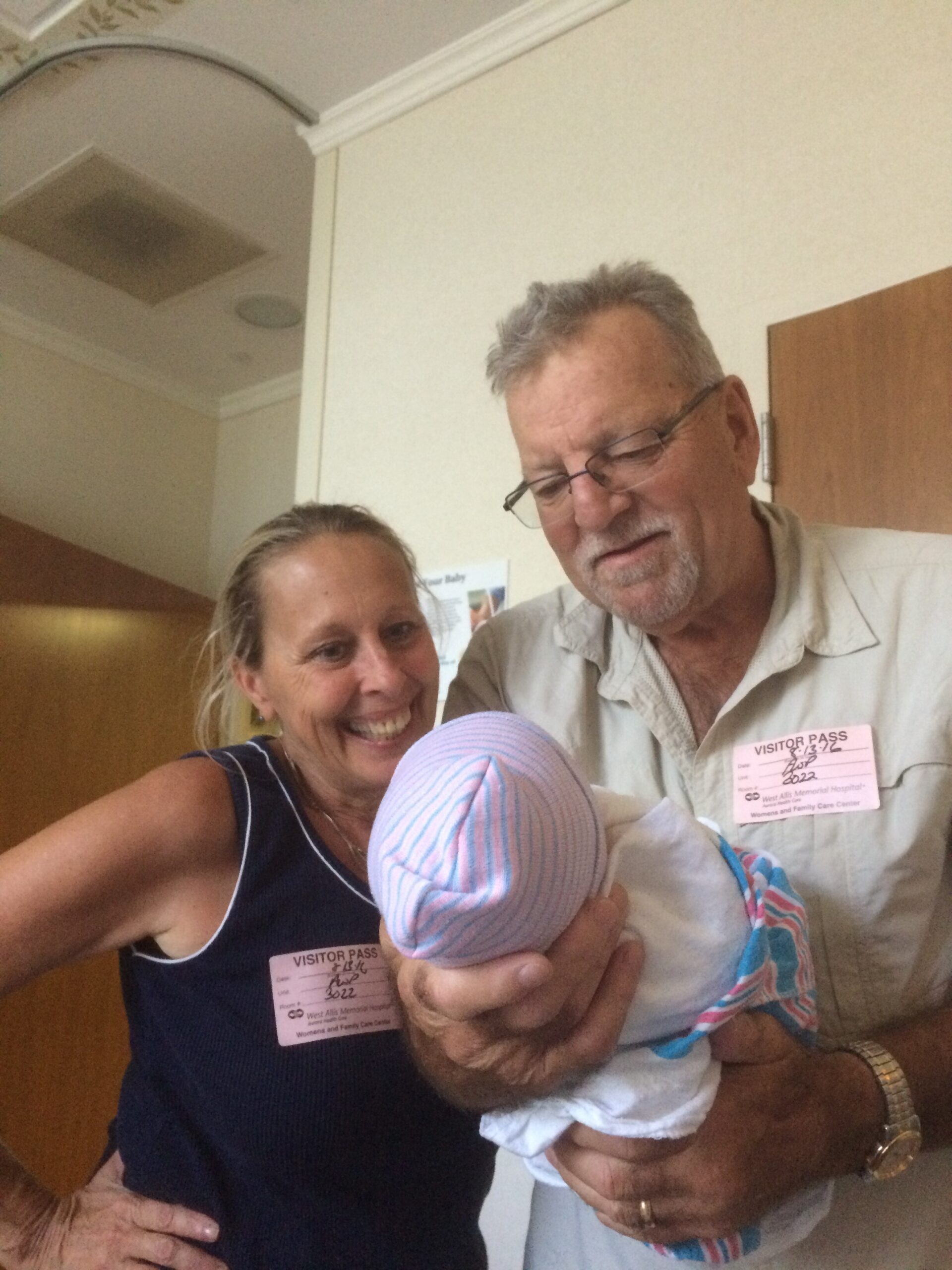 Smiling couple in a hospital hallway cradle a newborn baby wrapped in a striped blanket, both wearing visitor badges.