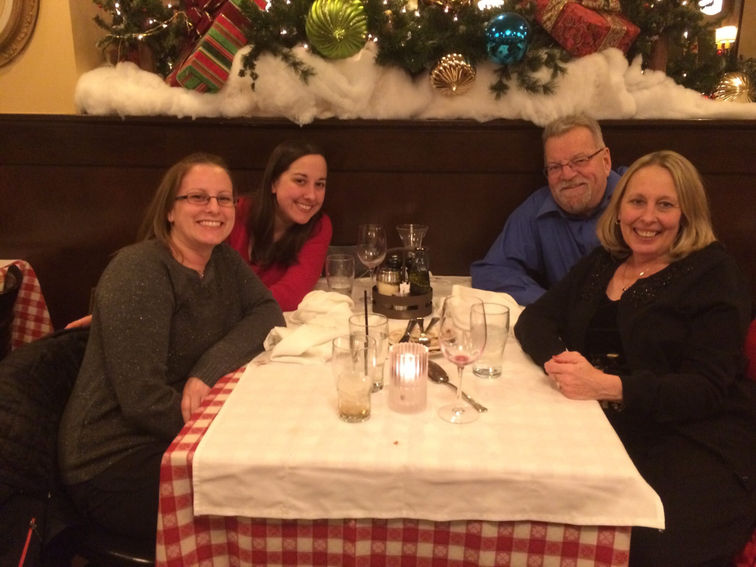 Four adults sit at a restaurant table smiling for the camera, holiday decor and a Christmas tree overhead in the background.