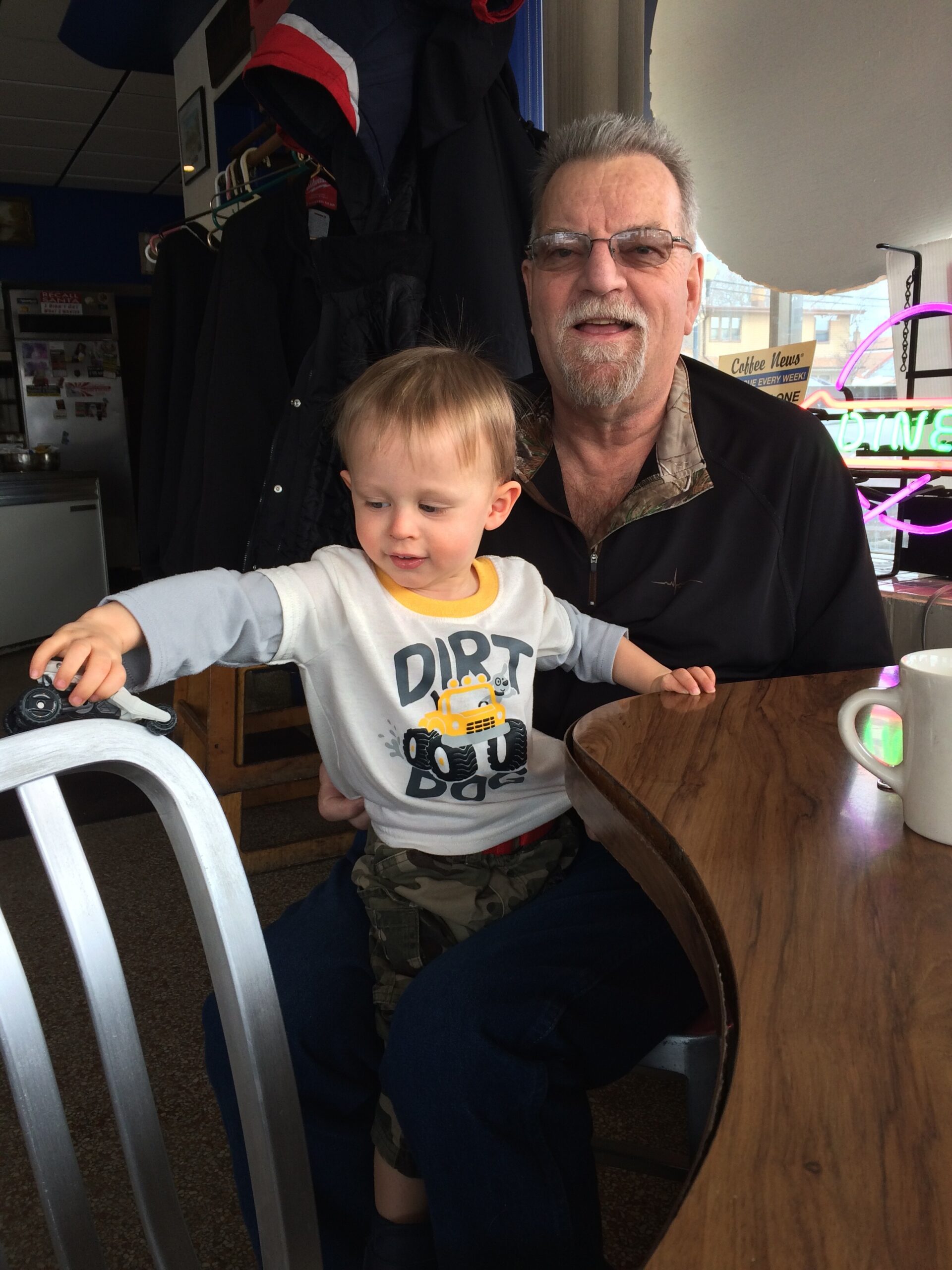Smiling older man with glasses holds a young boy at a cafe table; the child wears a 'Dirt Dogs' shirt and reaches for a toy car on the table.