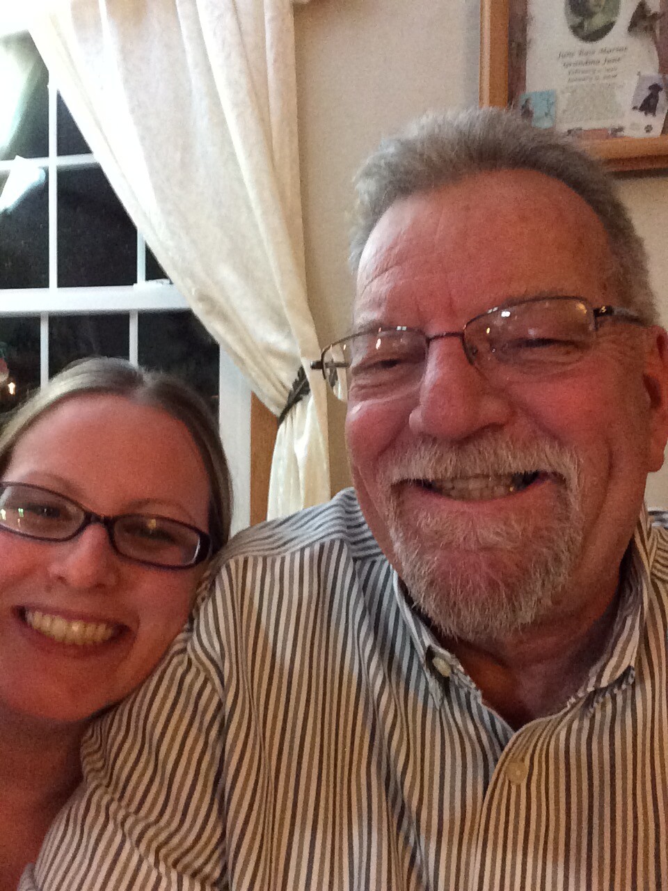 Two people smiling for a selfie indoors, a man with glasses and a striped shirt next to a woman with glasses and a bright smile in front of a window and curtains.