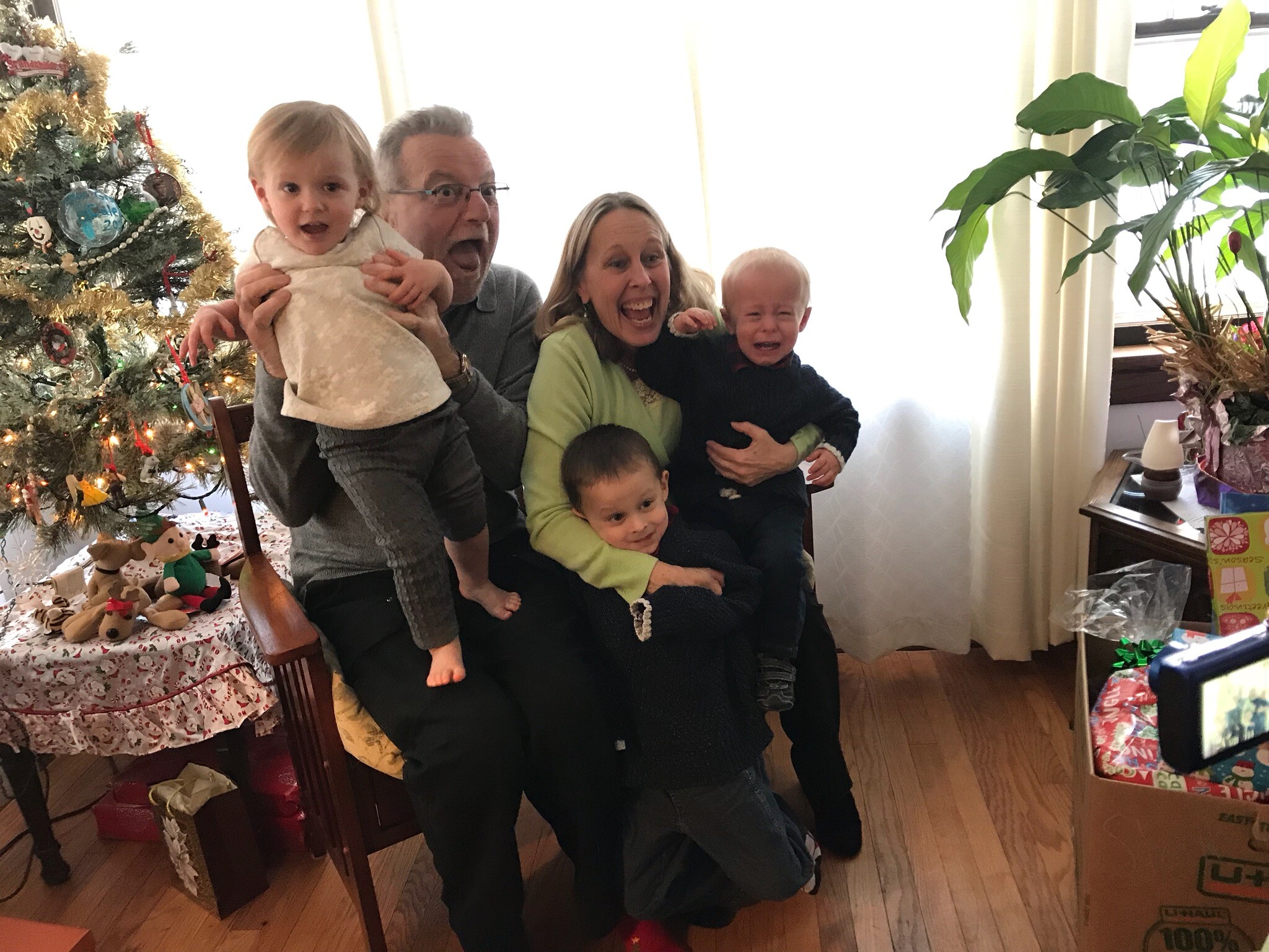 Family gathered for a Christmas photo: two adults and three young children posing by a decorated tree with presents nearby.