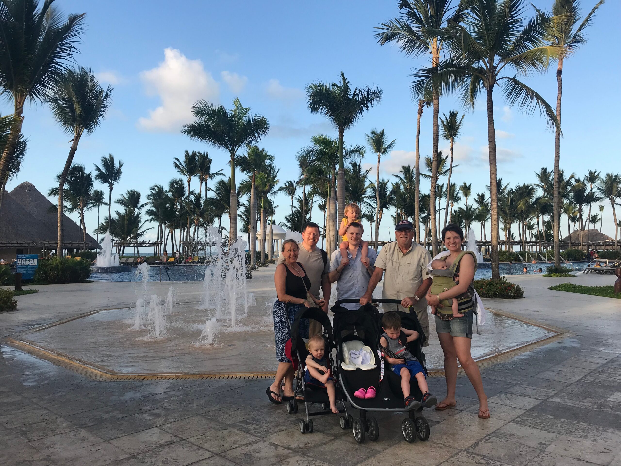 Family group posing together outdoors at a tropical resort with palm trees and a water fountain feature behind them.