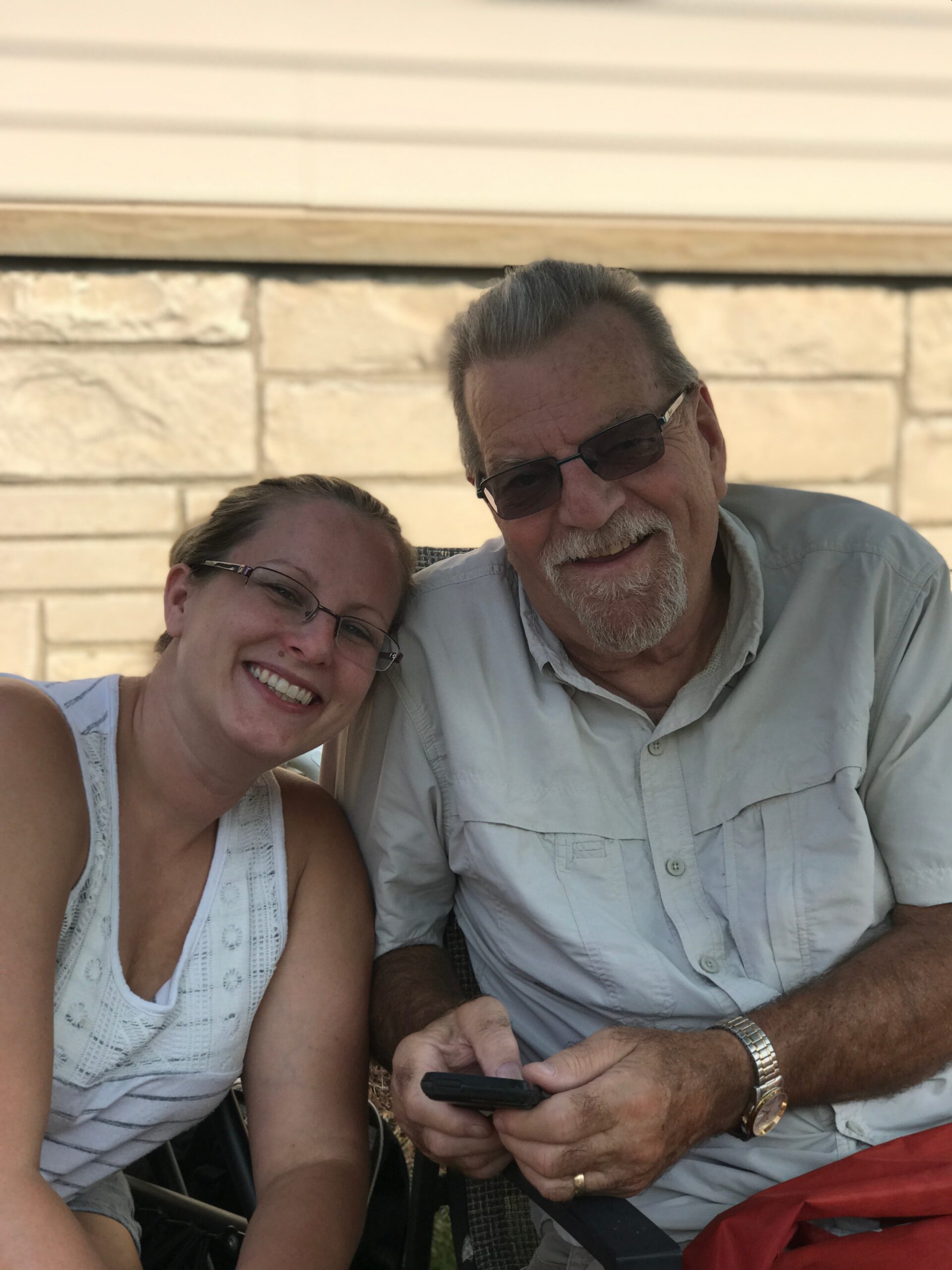 Smiling woman and older man wearing sunglasses sit together outdoors, man holding a smartphone.