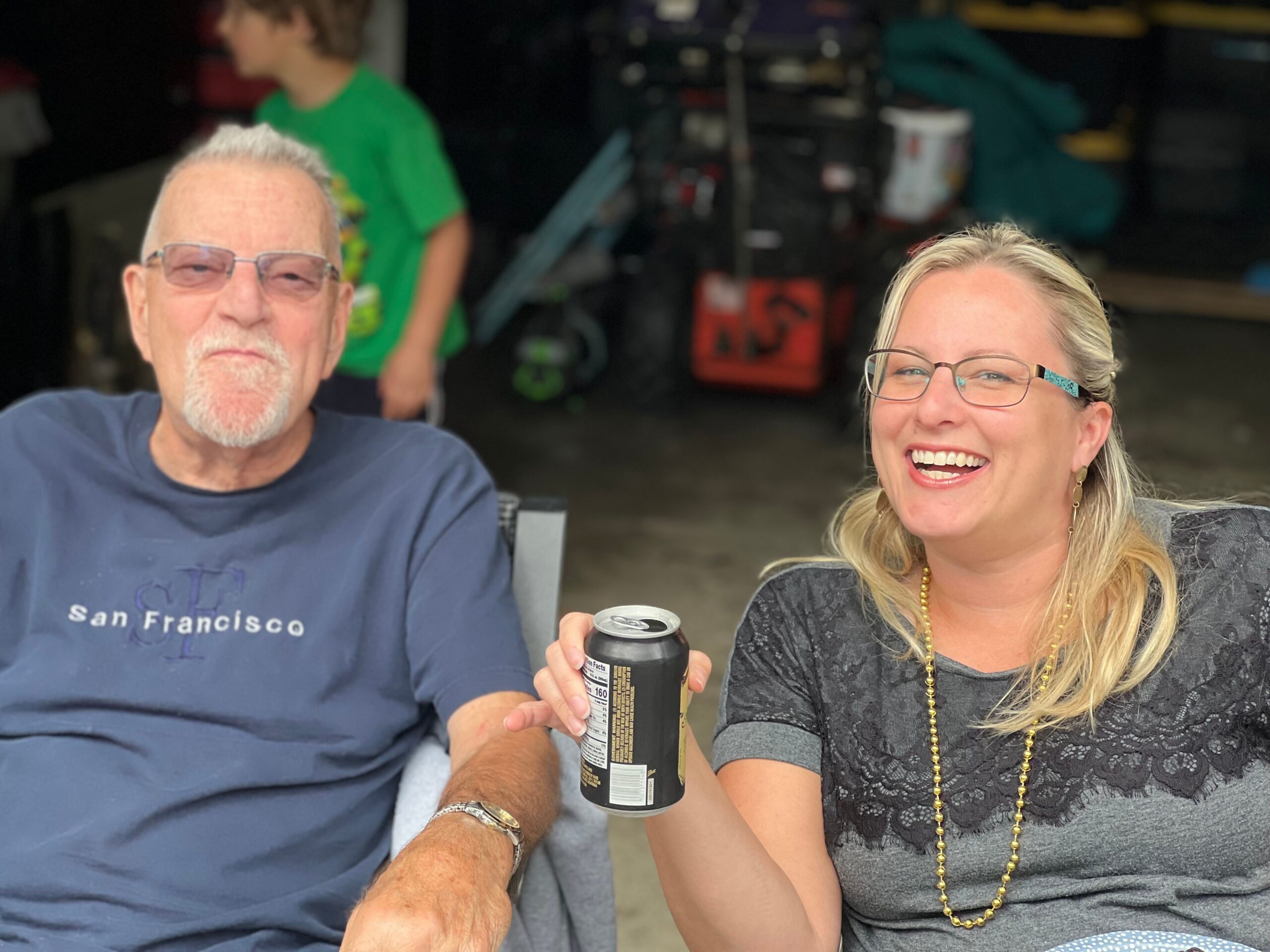 Two people smiling while sitting outdoors, each holding a can; a man in a blue shirt and a woman with glasses beside him, background shows a garage area with tools and items.