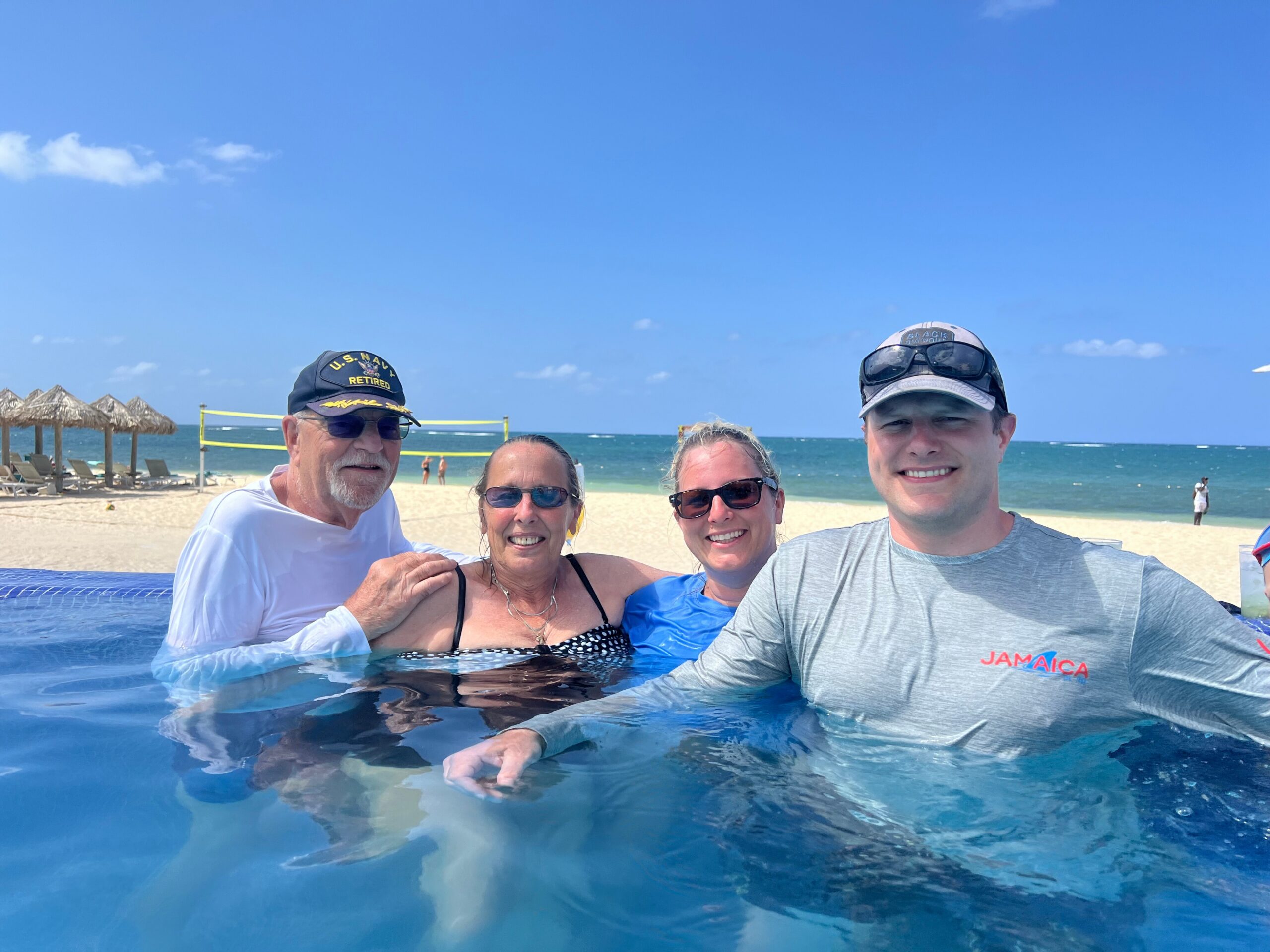 Group of four adults posing for a selfie in a shallow pool on a sunny beach, with blue sky, ocean, and beach umbrellas in the background. One person wears a Jamaica shirt.