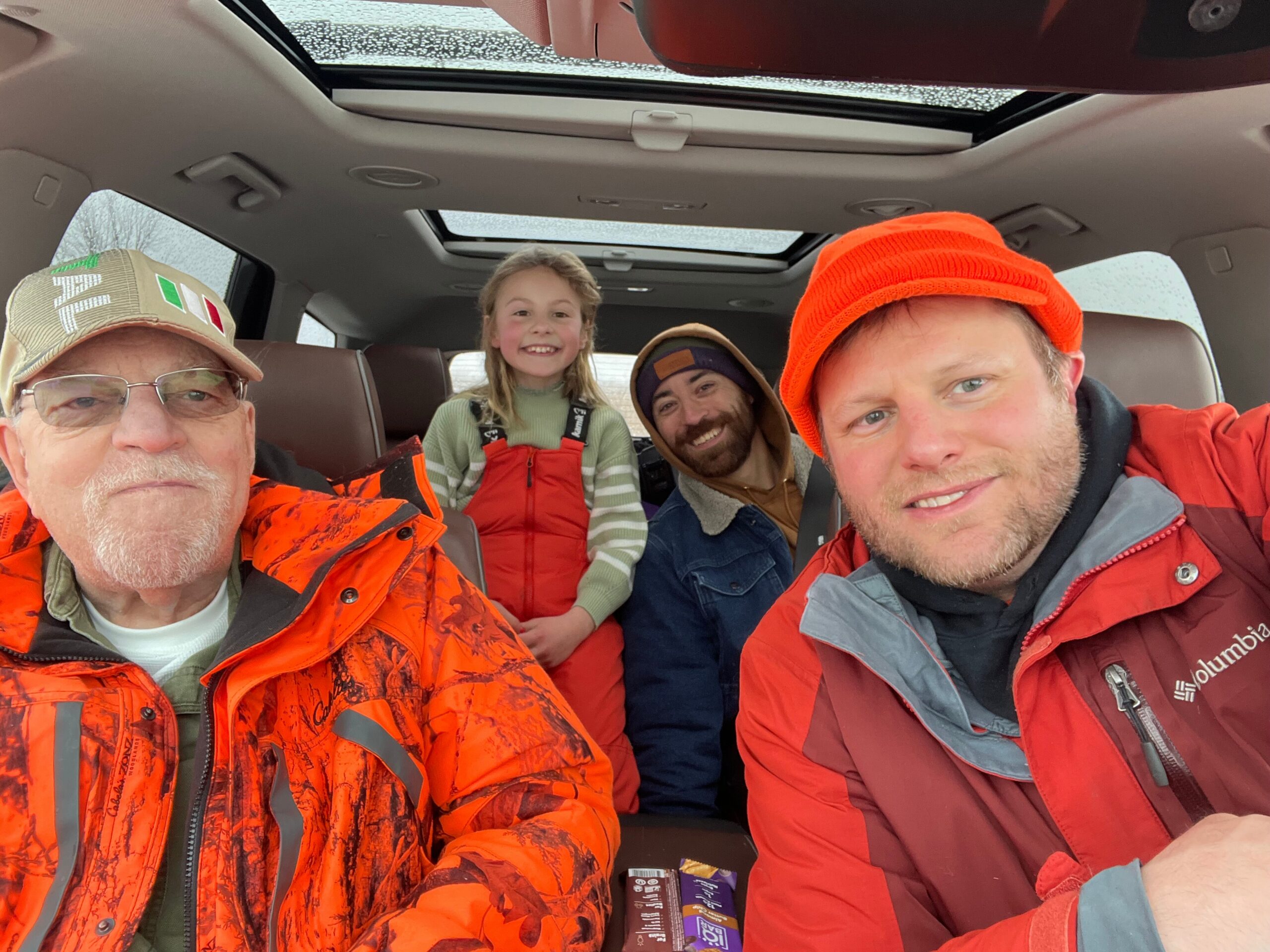 Four people wearing orange outerwear posing for a selfie inside a car with a sunroof, smiling at the camera.