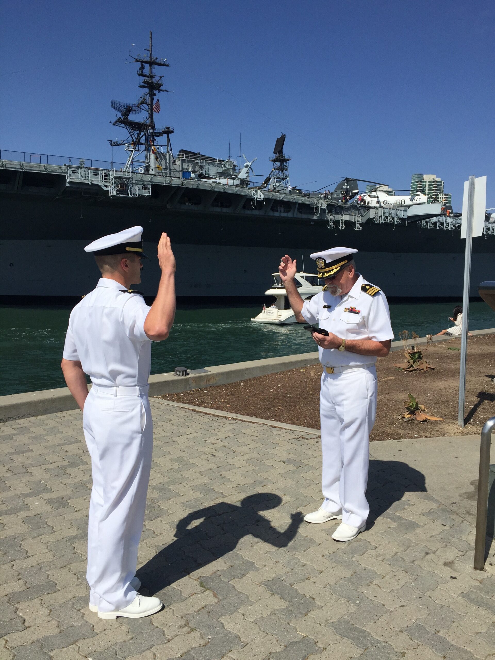 Two Navy officers in white uniforms salute on a riverside promenade with a large aircraft carrier in the background.
