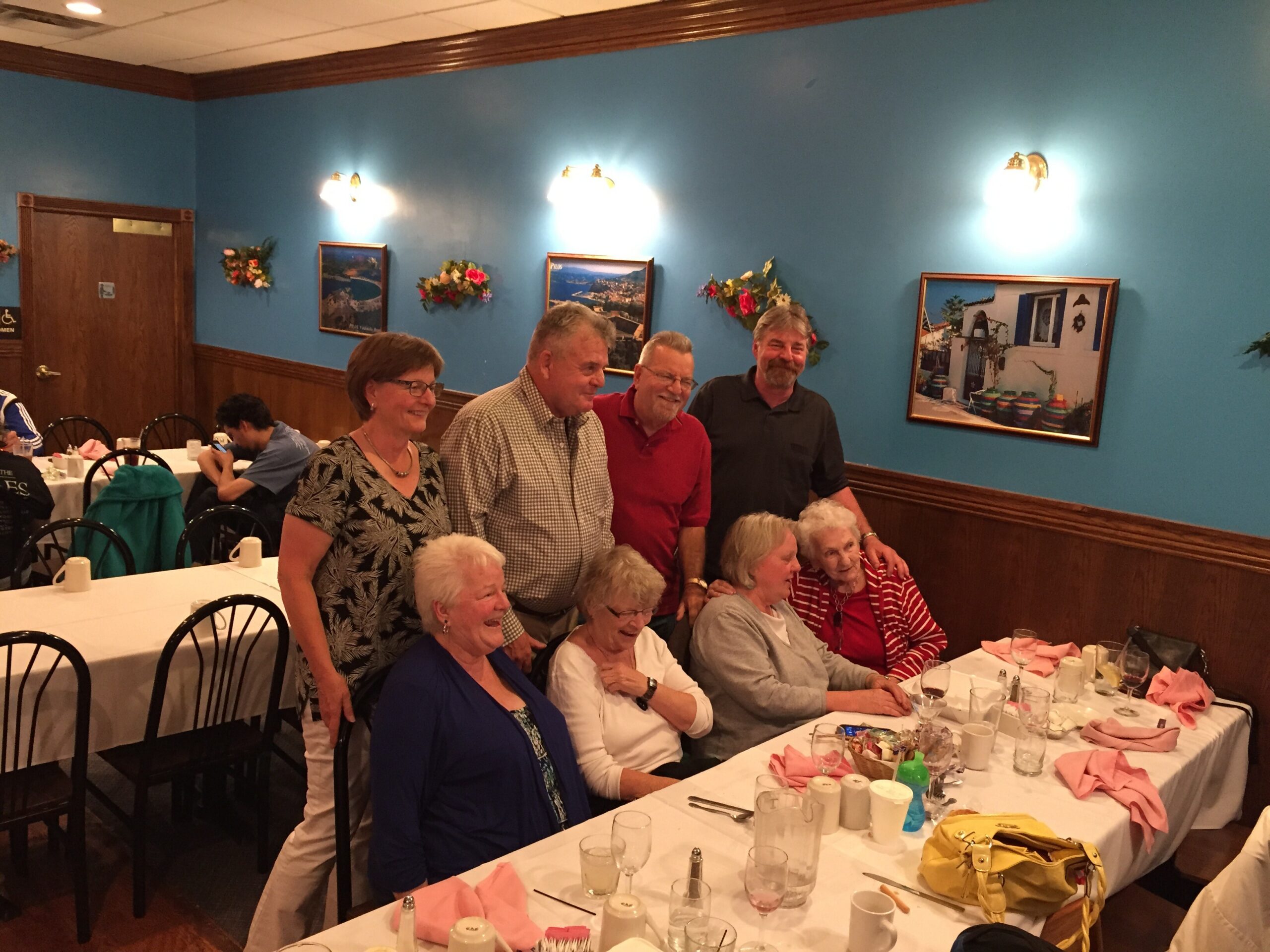 Group of seniors smiling at a long restaurant table with pink napkins and dishes on white tablecloths against a blue wall.
