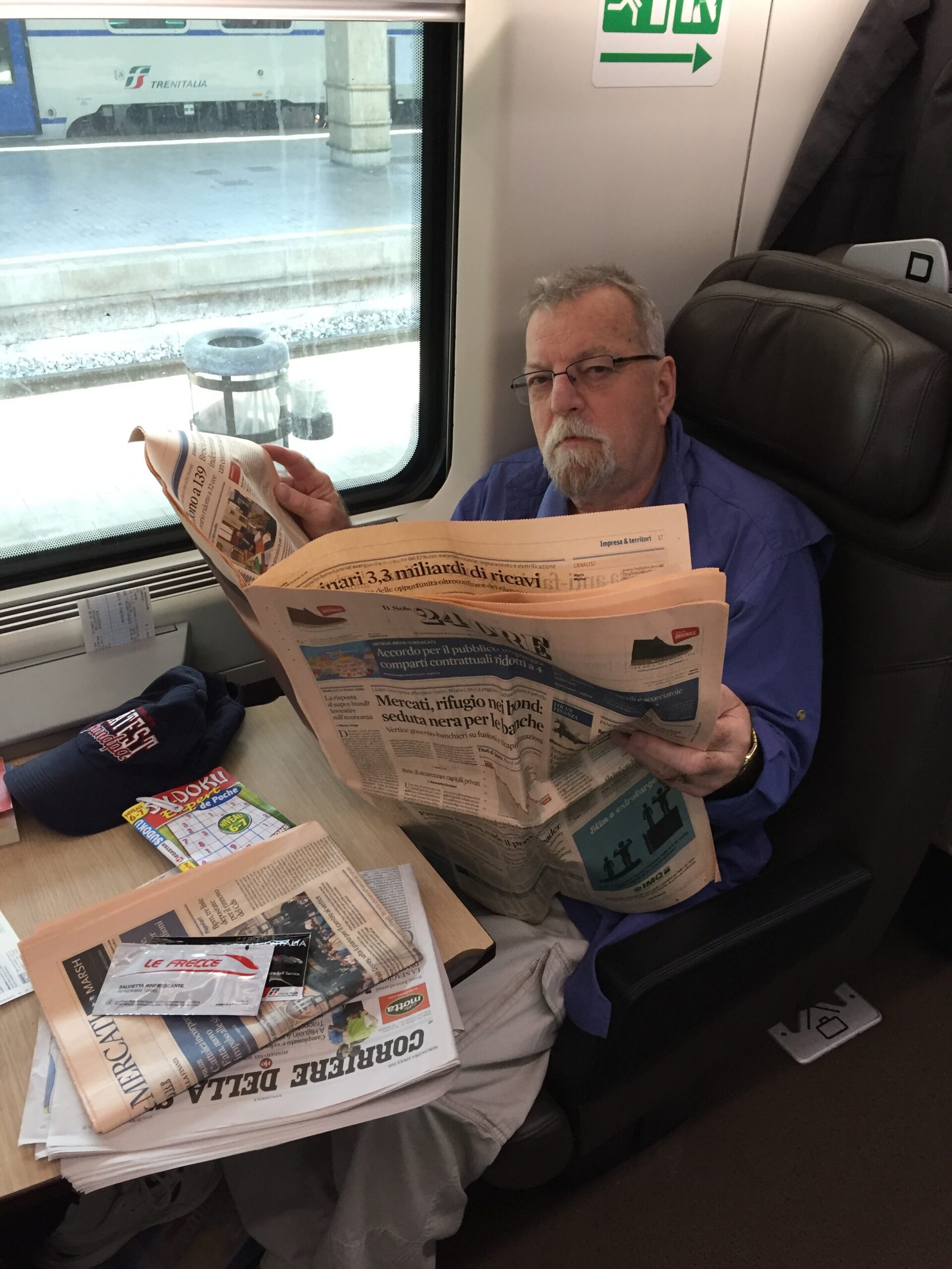 Man in a blue shirt sits on a train seat, reading a large Italian newspaper at a window-side table, with more papers spread across the table and a baton-like trash can visible outside the window.