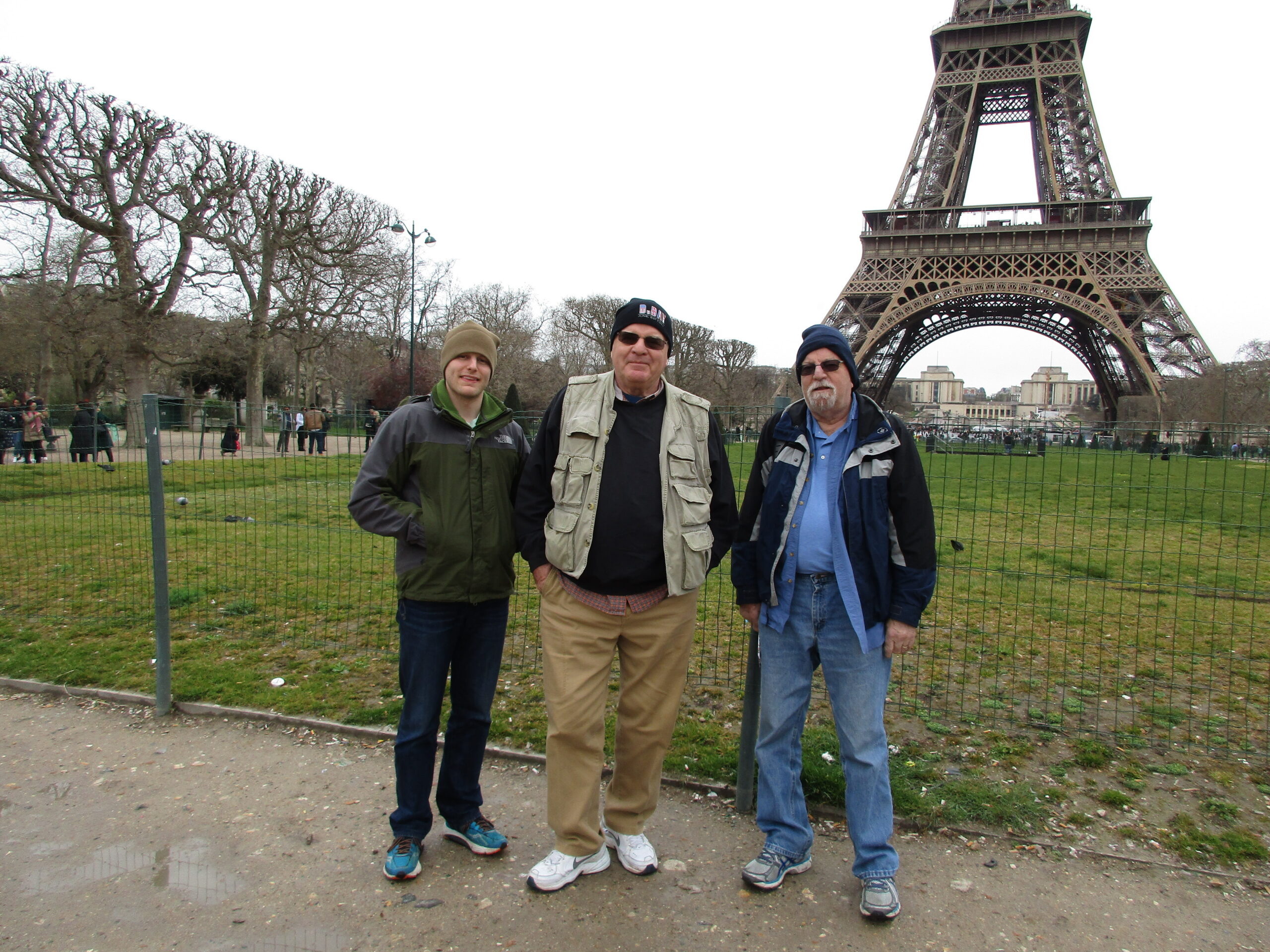 Three men posing for a photo in a park with the Eiffel Tower in the background, fenced area and leafless trees nearby.