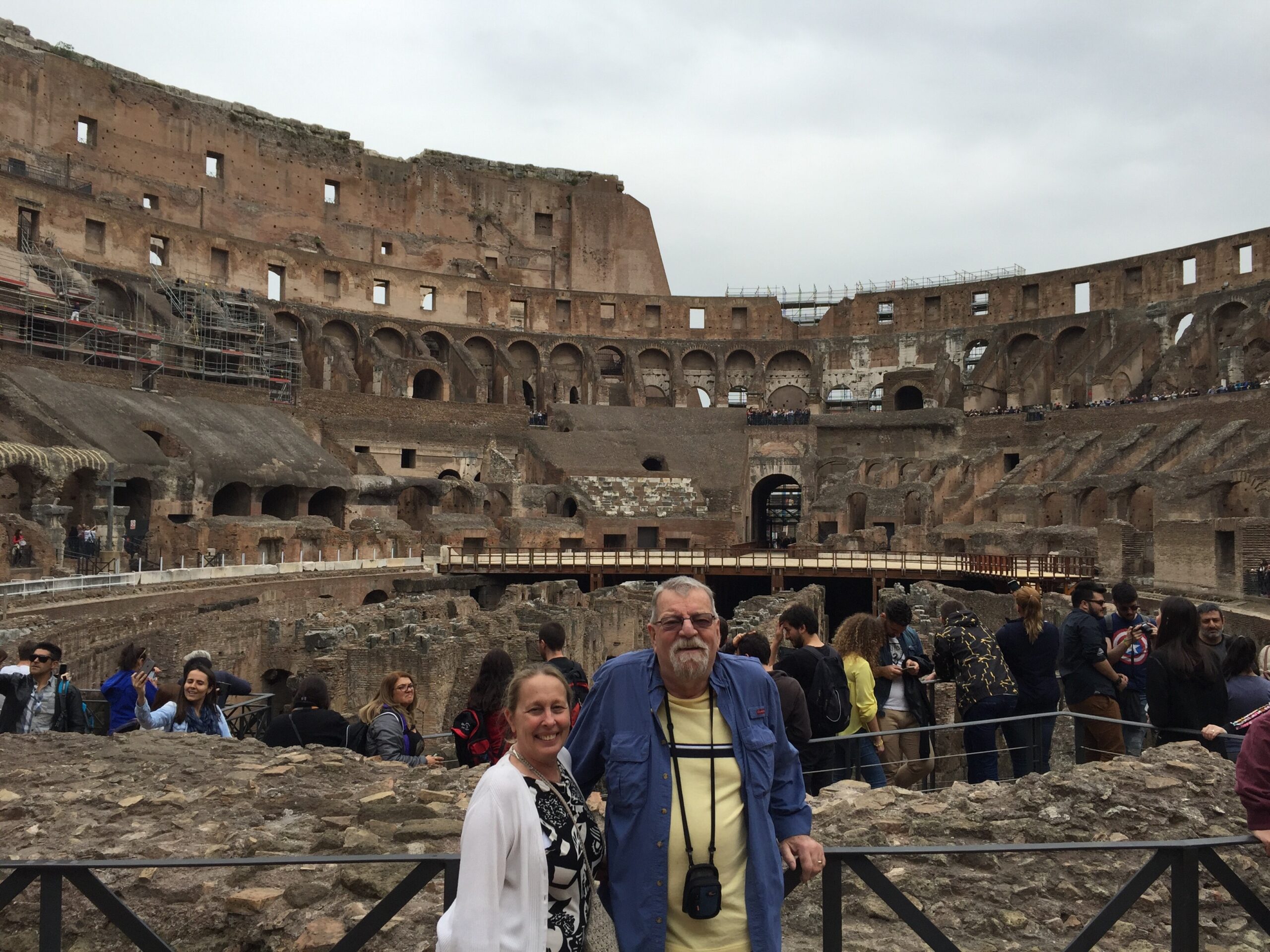 Tourists stand inside the ancient Roman Colosseum, with arched walls and scaffolding on the left against an overcast sky.