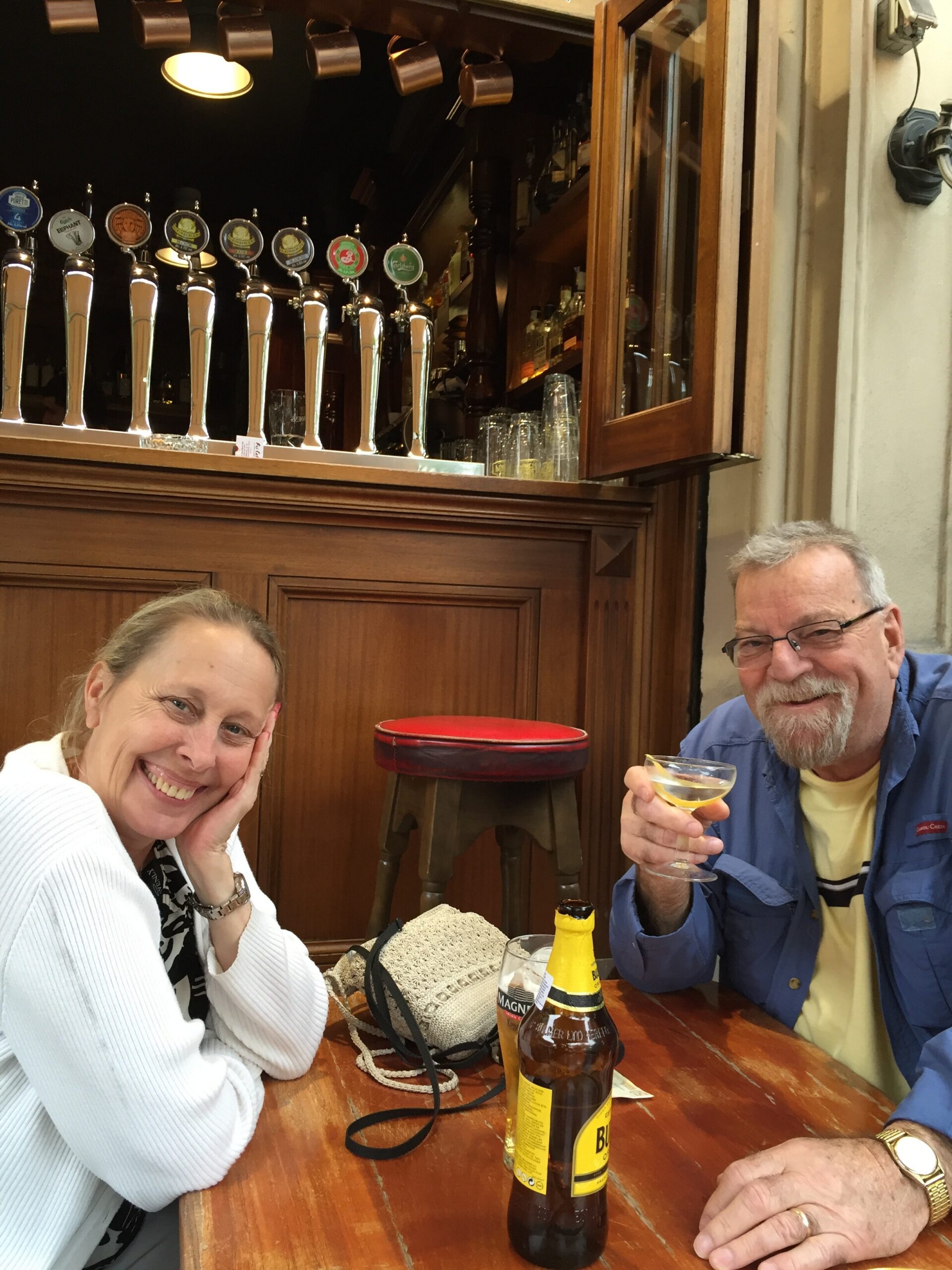 Smiling woman and man sit at a wooden pub table with beer taps behind them; the man holds a glass of cider and a Magners bottle is on the table.