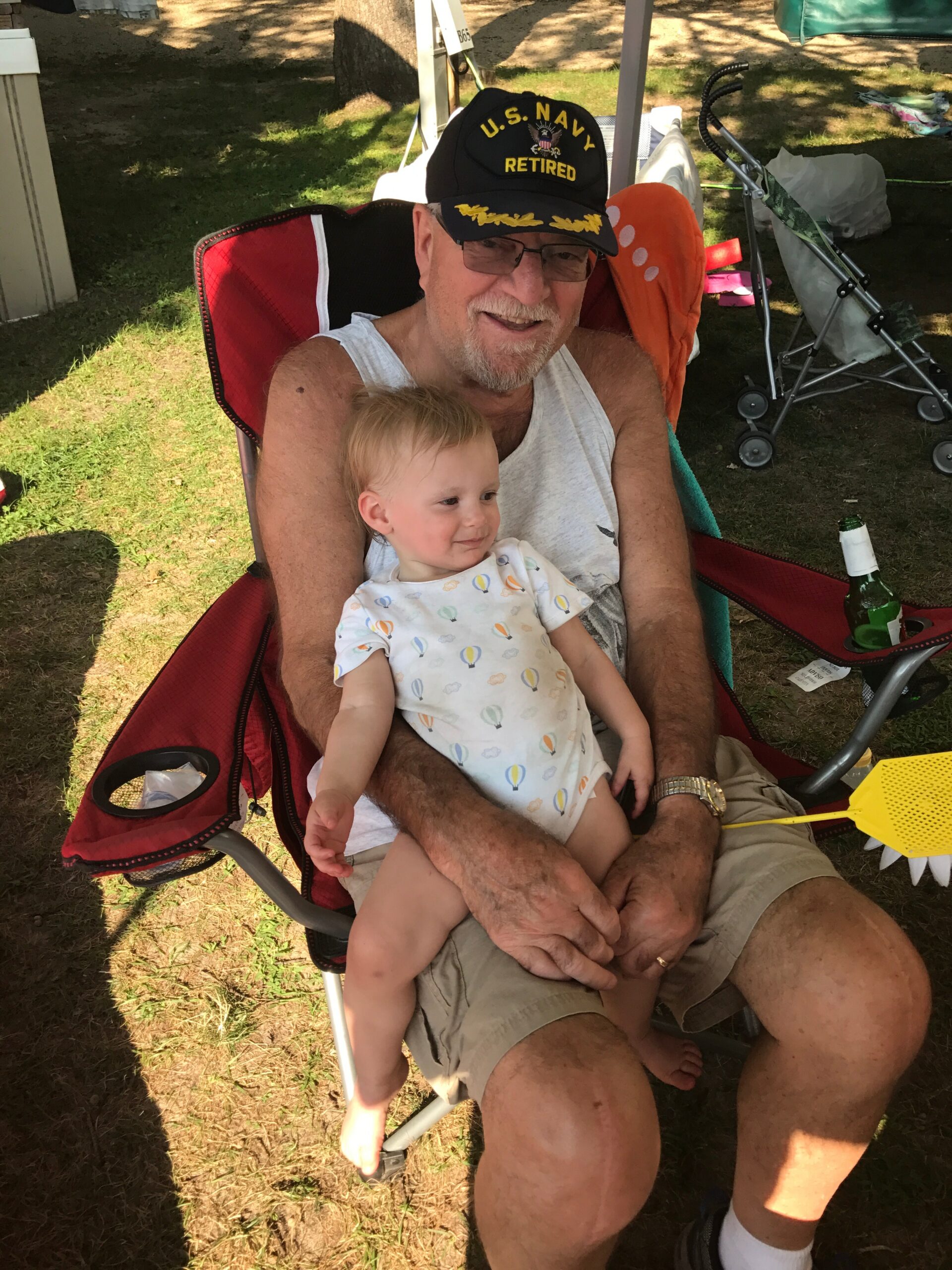Older man wearing a U.S. Navy Retired cap sits in a red camping chair outdoors, holding a smiling baby on his lap during a sunny day in the park.