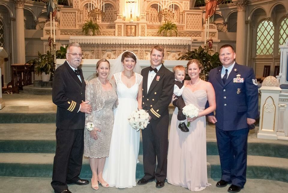 Wedding party posing inside an ornate church: bride in white gown, groom in uniform, and two parents with a young child nearby.