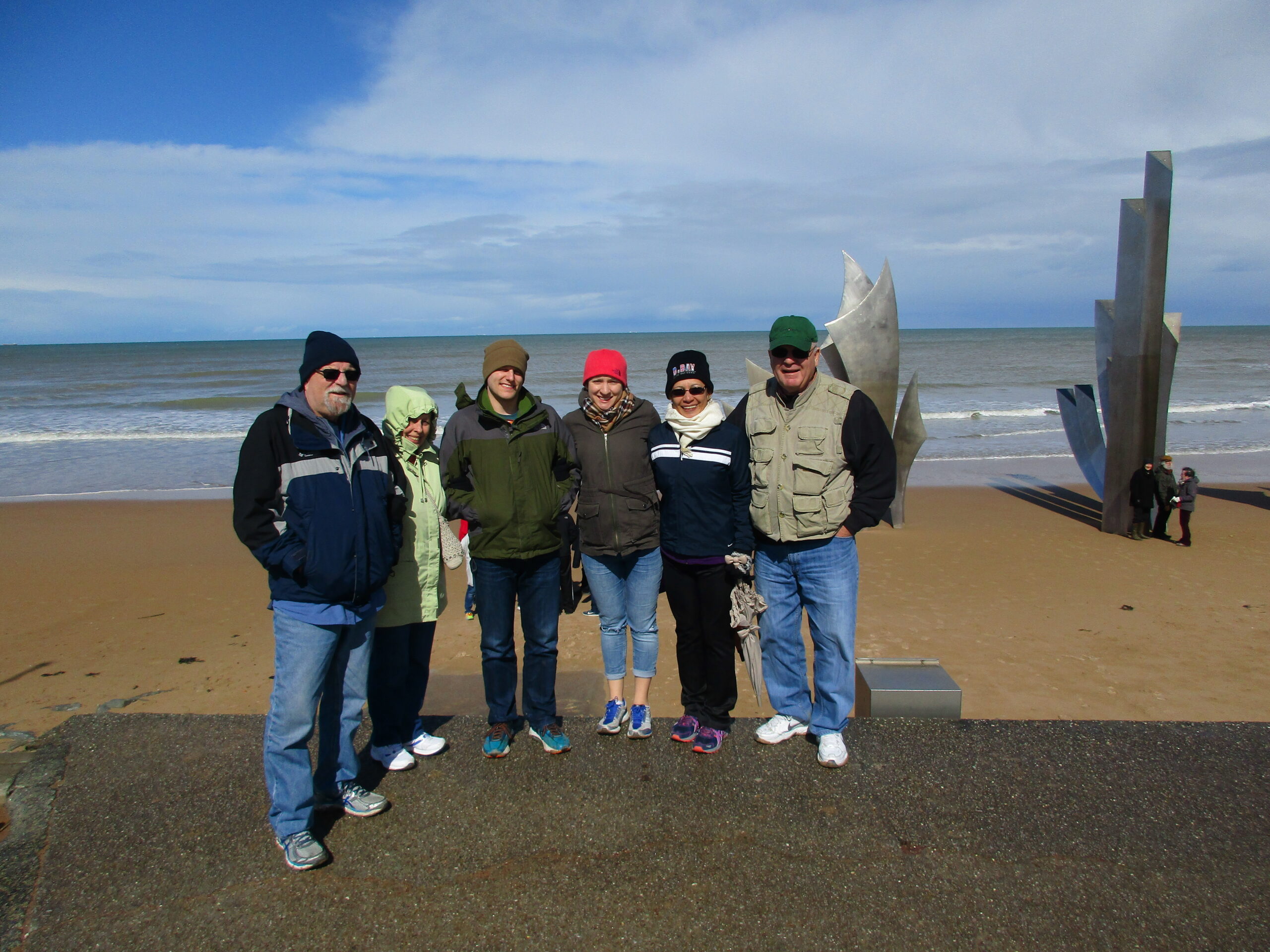 A group of adults posing on a sandy beach with a large abstract metal sculpture in the background under a blue sky and clouds.