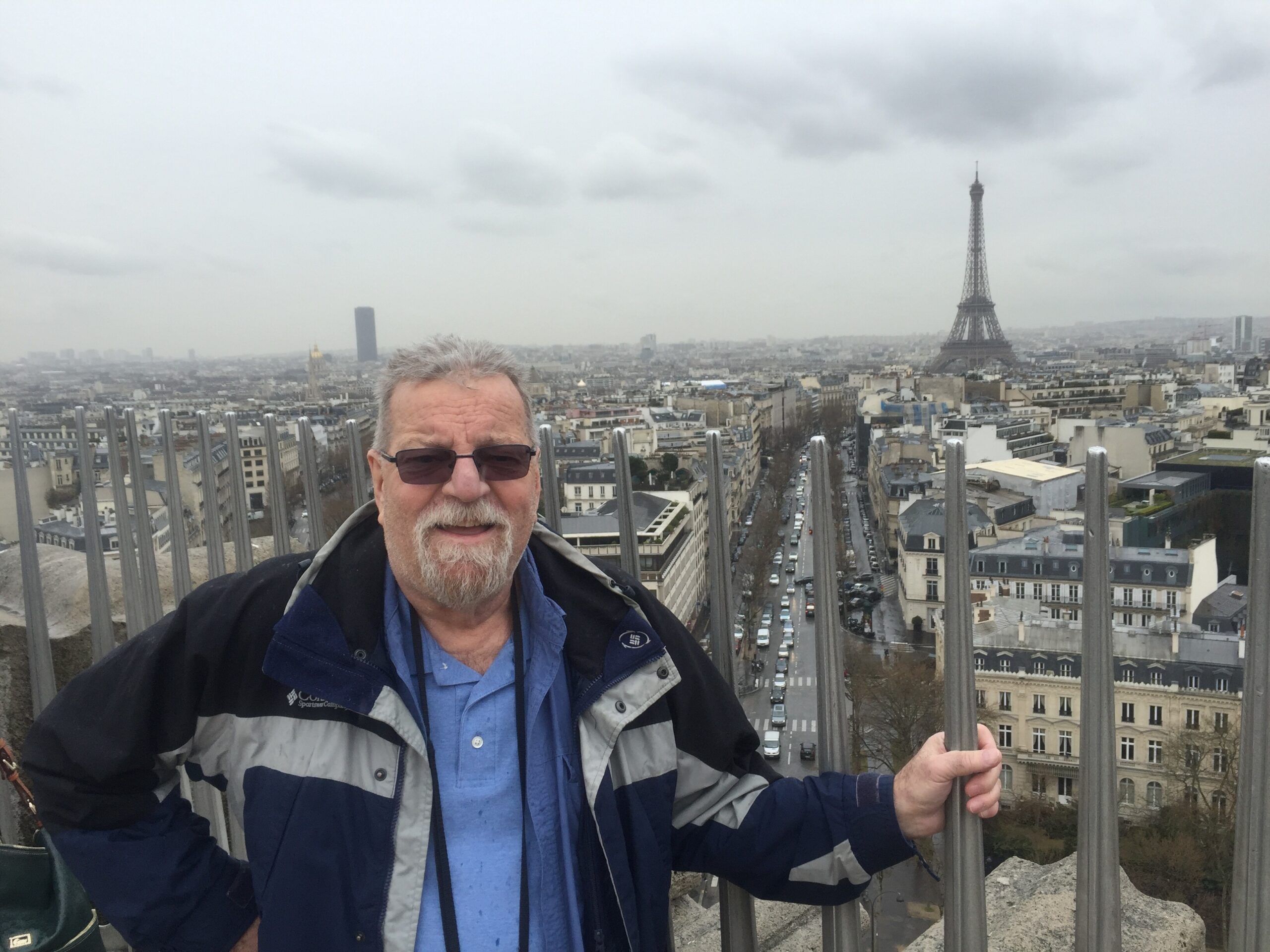 Selfie of a man in a blue shirt and navy jacket on a rooftop railing with Paris city skyline and the Eiffel Tower in the distance