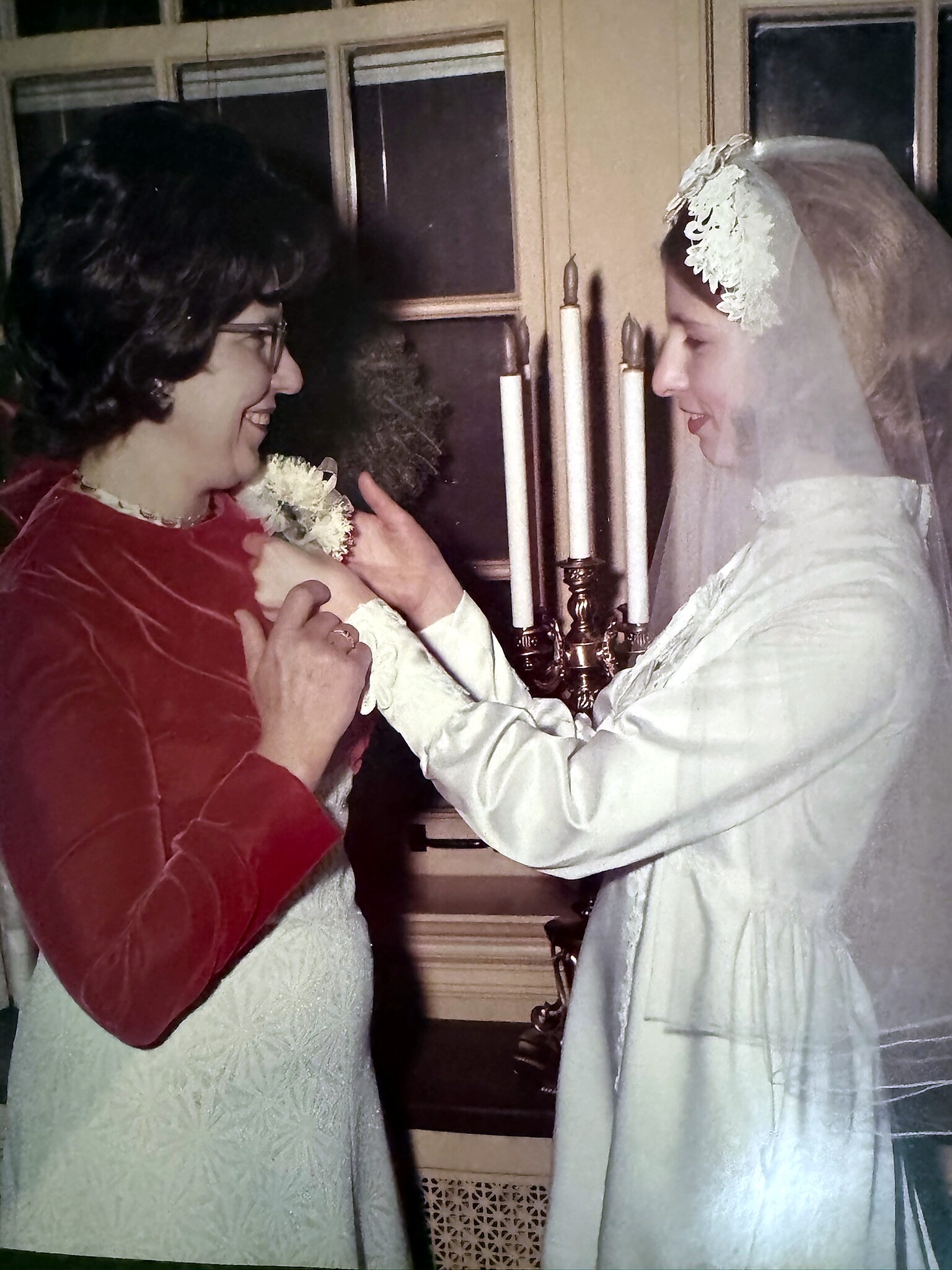 Bride in a white gown and lace veil smiles at a woman in a red velvet cape as they touch shoulders beside a candle holder.