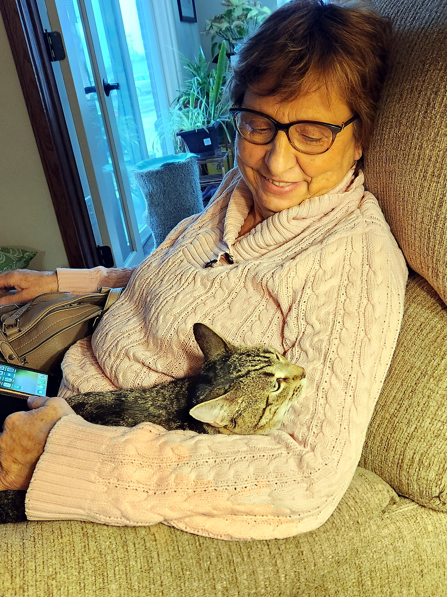 Older woman with short hair and glasses, wearing a pink cable-knit sweater, sits on a sofa with a tabby cat on her lap, smiling contentedly.