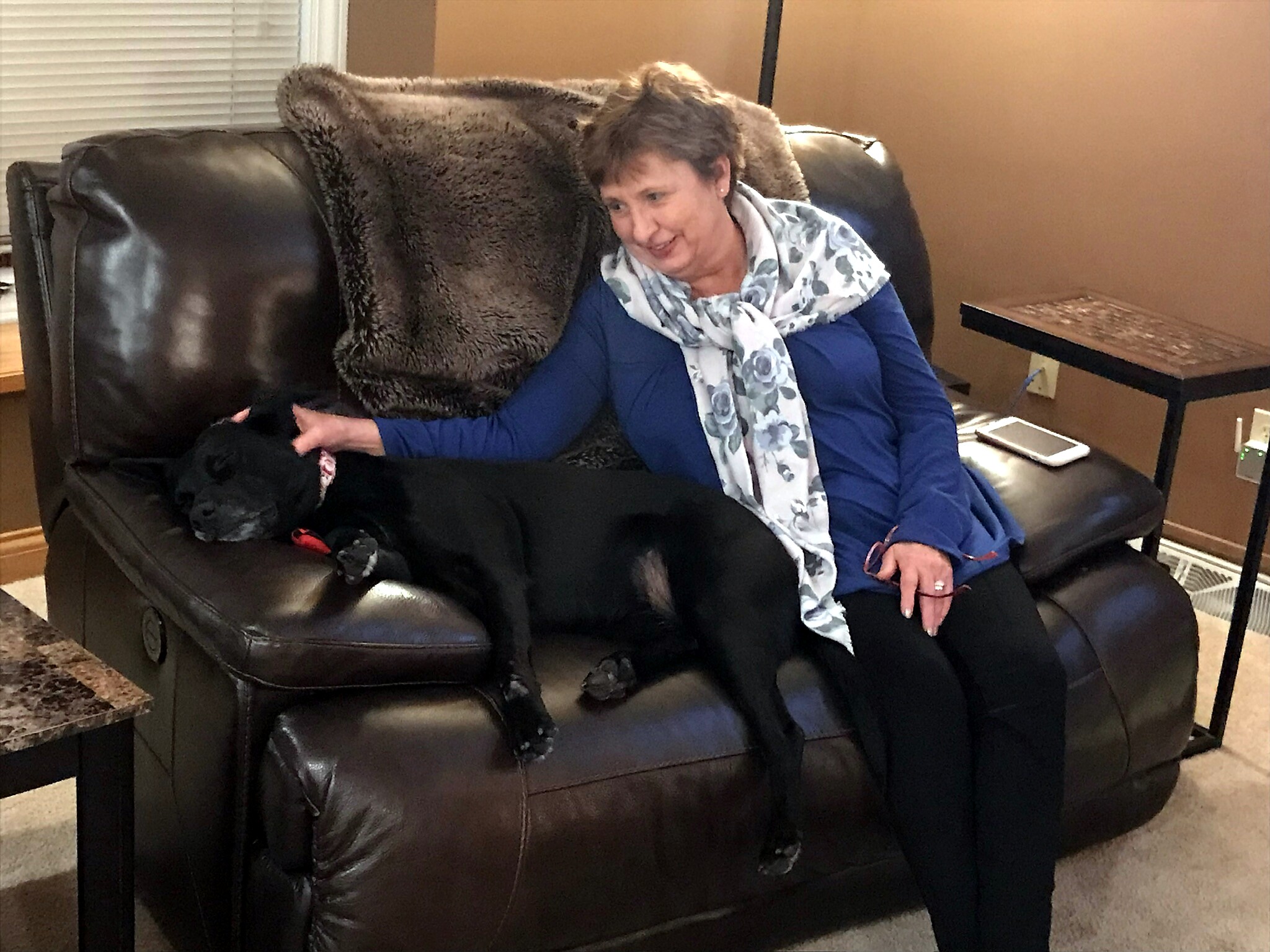 Woman in a blue top and floral scarf sits on a dark leather sofa, smiling, with a large black dog resting beside her on the couch and a brown blanket draped over the back.