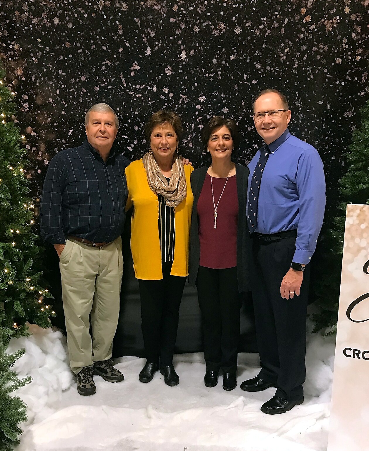 Four adults stand together indoors in front of a snowy black backdrop with Christmas trees on the sides, smiling at the camera.