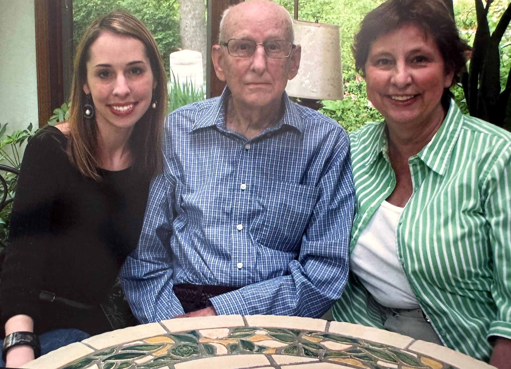 Three adults pose at an outdoor garden table with a mosaic top, smiling as greenery surrounds them.