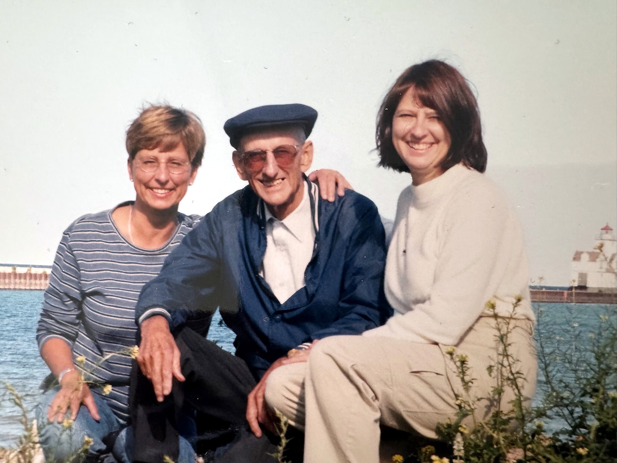 Three people posing outdoors by the water, smiling at the camera; man in the center wears a blue cap, flanked by two women.